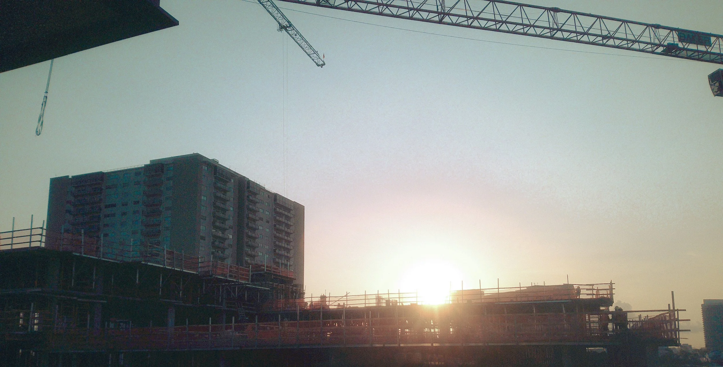 Construction site with a crane, building framework, and a tall residential building under construction at sunrise.