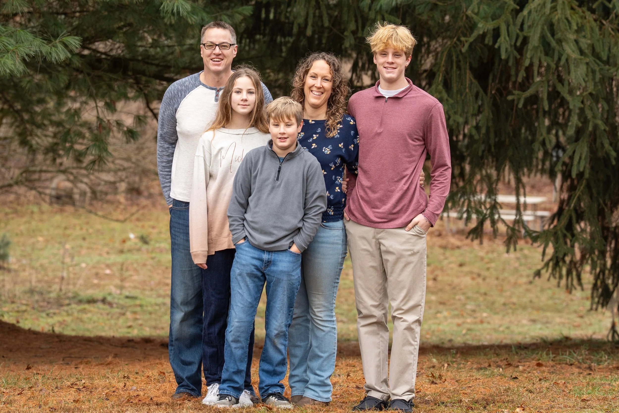 A family of six standing outdoors beneath pine trees, smiling and enjoying a casual moment.