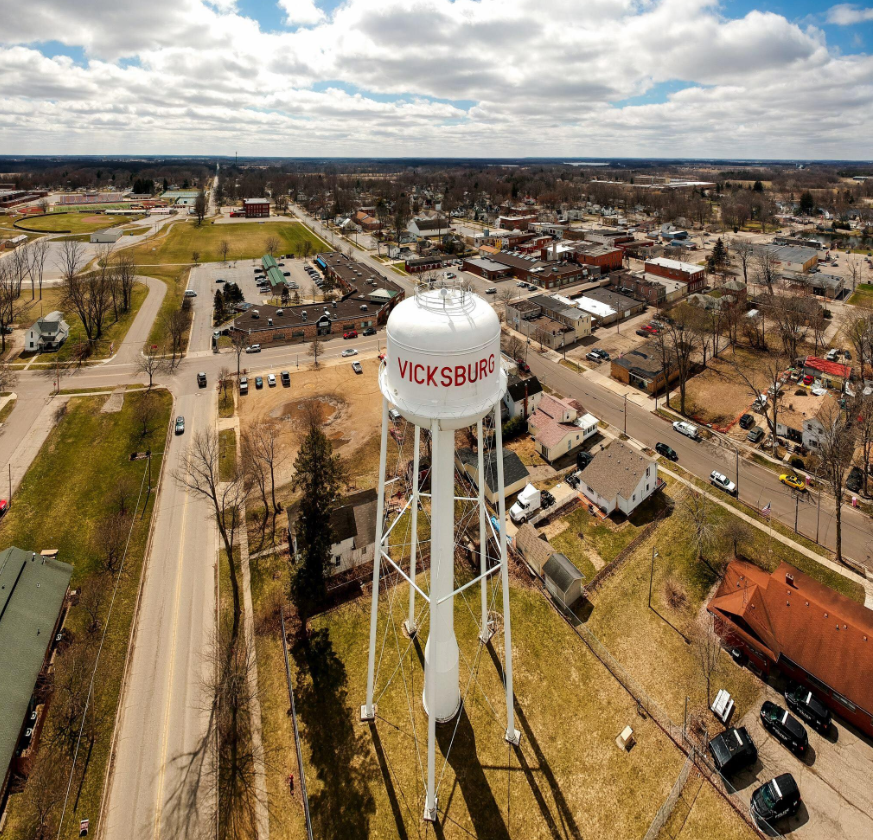 Aerial view of Vicksburg water tower surrounded by residential and commercial buildings, streets, and a park with sports fields under partly cloudy skies. To symbolize community Alignment and trust.