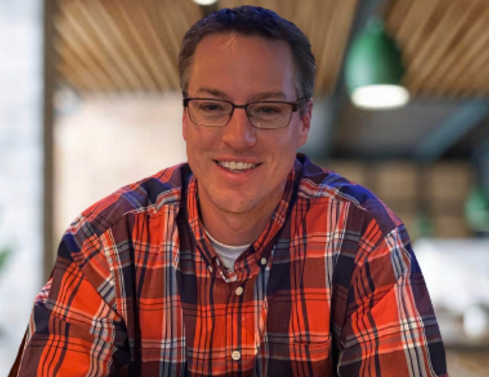 A young man with glasses smiling, wearing a red and blue plaid shirt, indoors with wooden ceiling and green lamps.