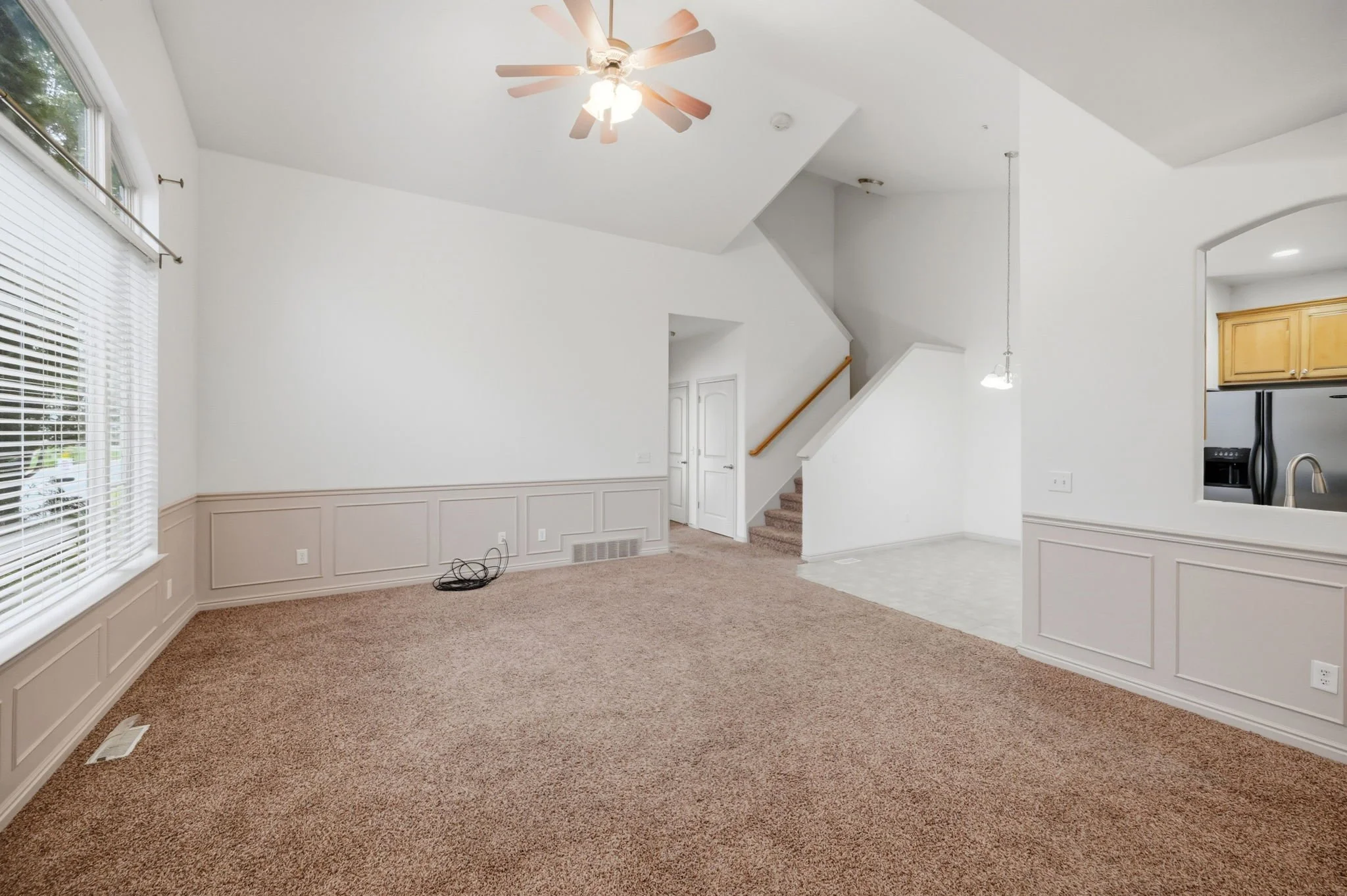 Empty living room with beige carpeting, white walls with chair molding, large window with blinds, ceiling fan, staircase, and an open kitchen area.