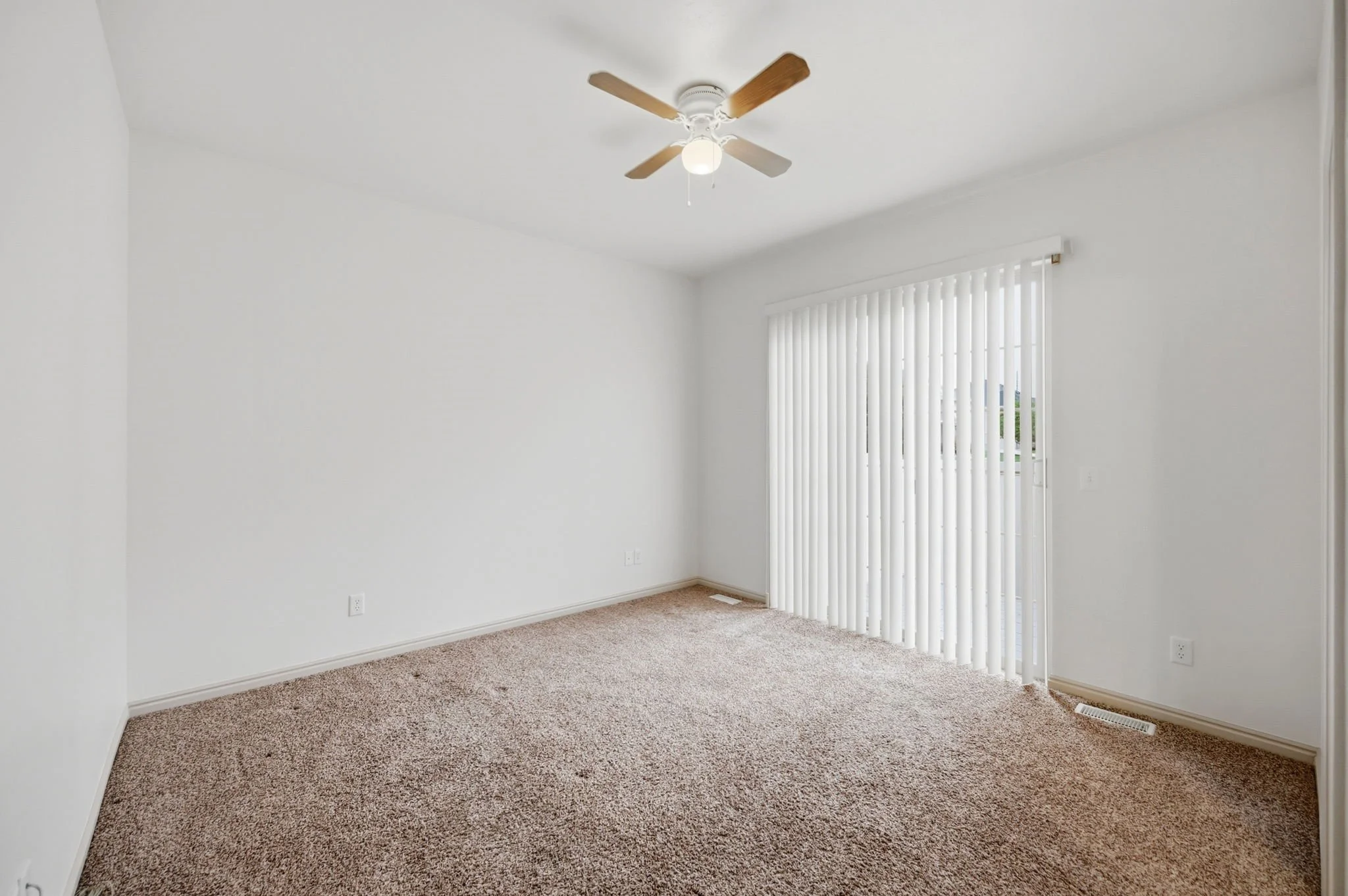 Empty room with beige carpet, white walls, a ceiling fan with wooden blades, vertical blinds covering a window, and a heating vent on the floor.