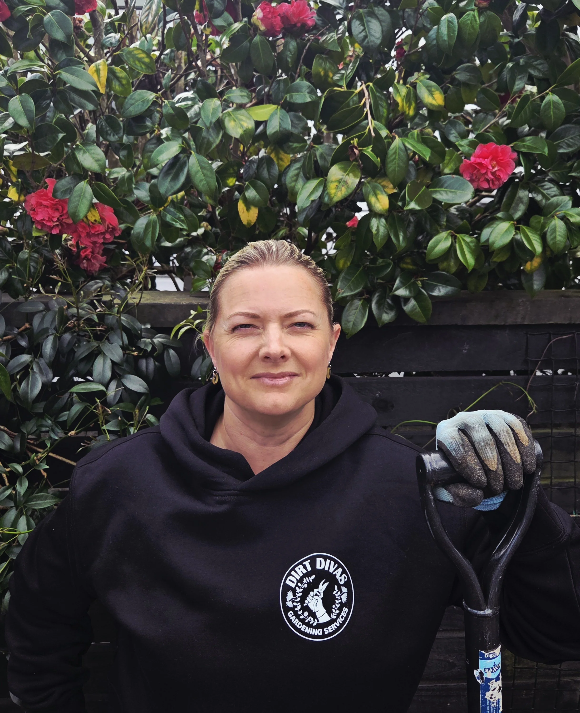 Woman with light skin, blonde hair tied back, wearing a black sweatshirt with a logo that reads "Dirt Divas Gardening Services," holding a gloved hand and standing outdoors in front of leafy shrubs with pink flowers.