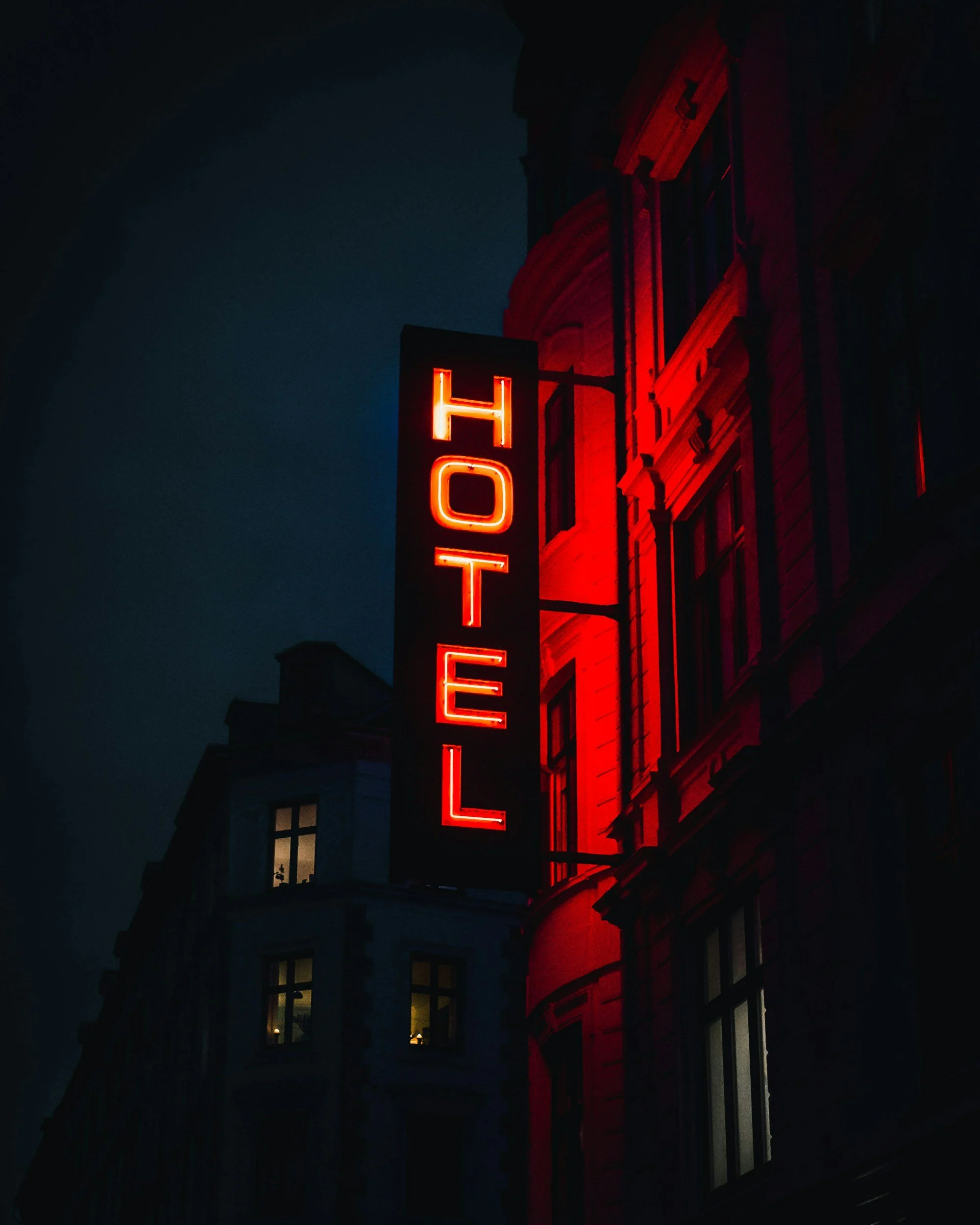 Nighttime photo of a building with a vertical red neon sign reading 'HOTEL' in capital letters, illuminated against a dark sky, with windows visible on the building.