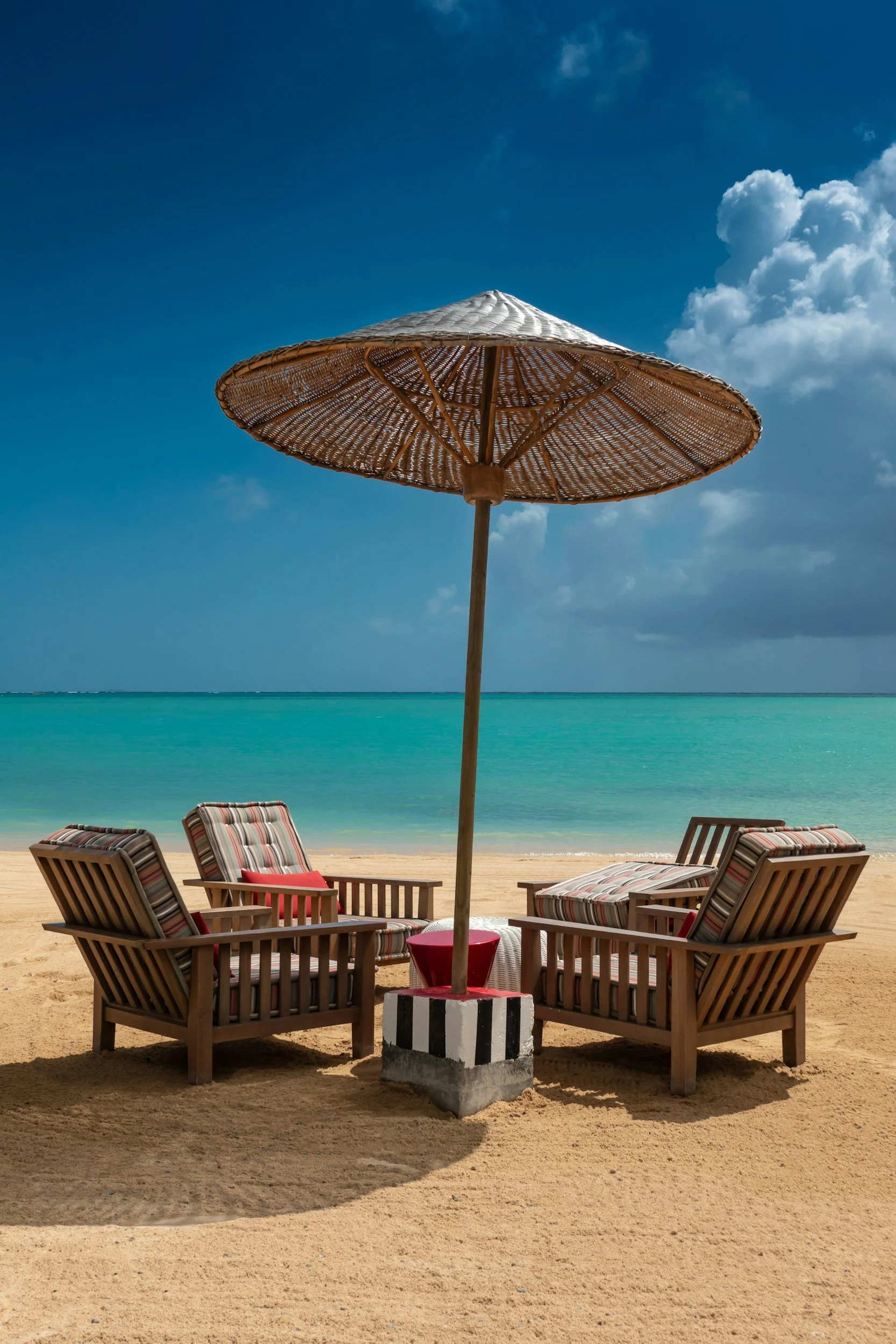 A beach scene with four wooden lounge chairs with striped cushions arranged in a circle, under a large straw sun umbrella. The ocean and blue sky with some clouds are visible in the background.