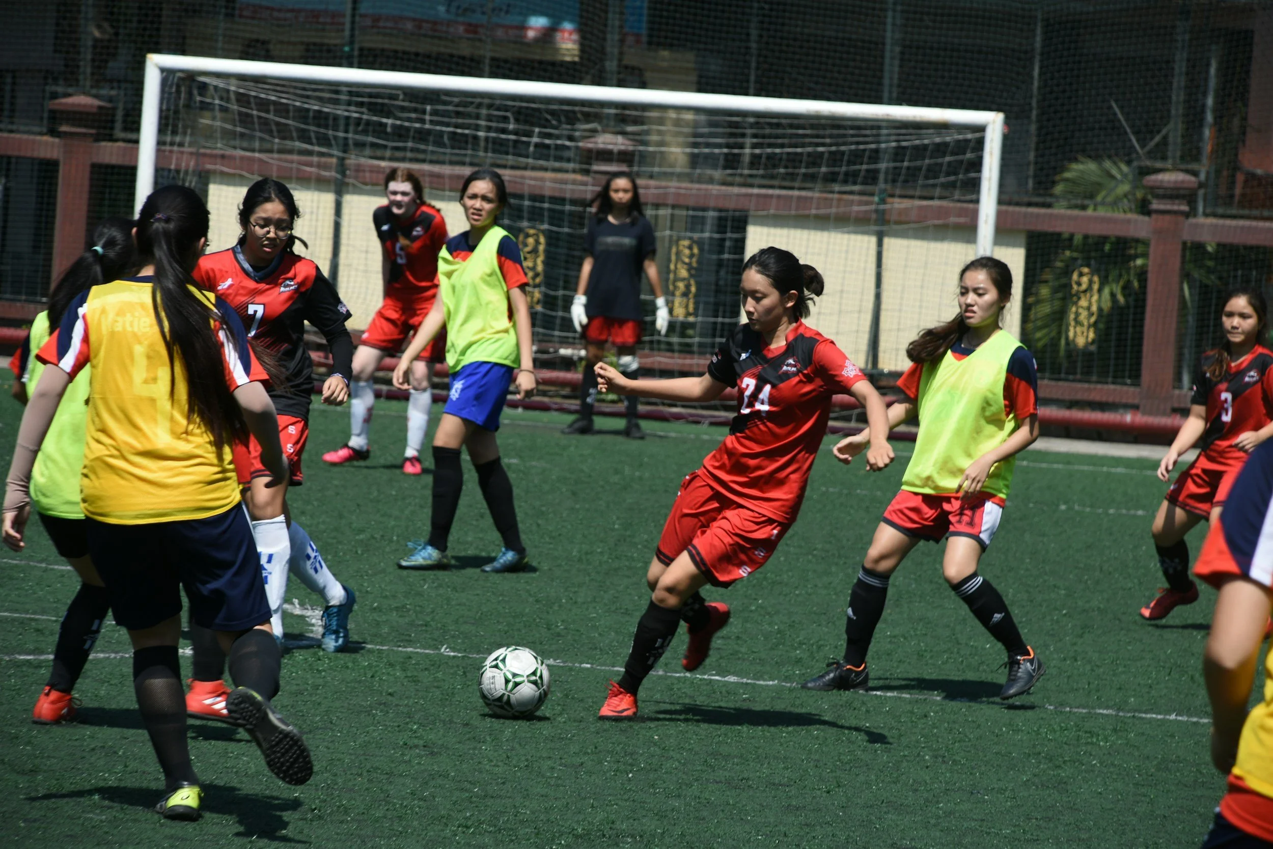 Young girls playing soccer on a field, with some wearing yellow bibs and others in red uniforms, near a goalpost in an outdoor setting.