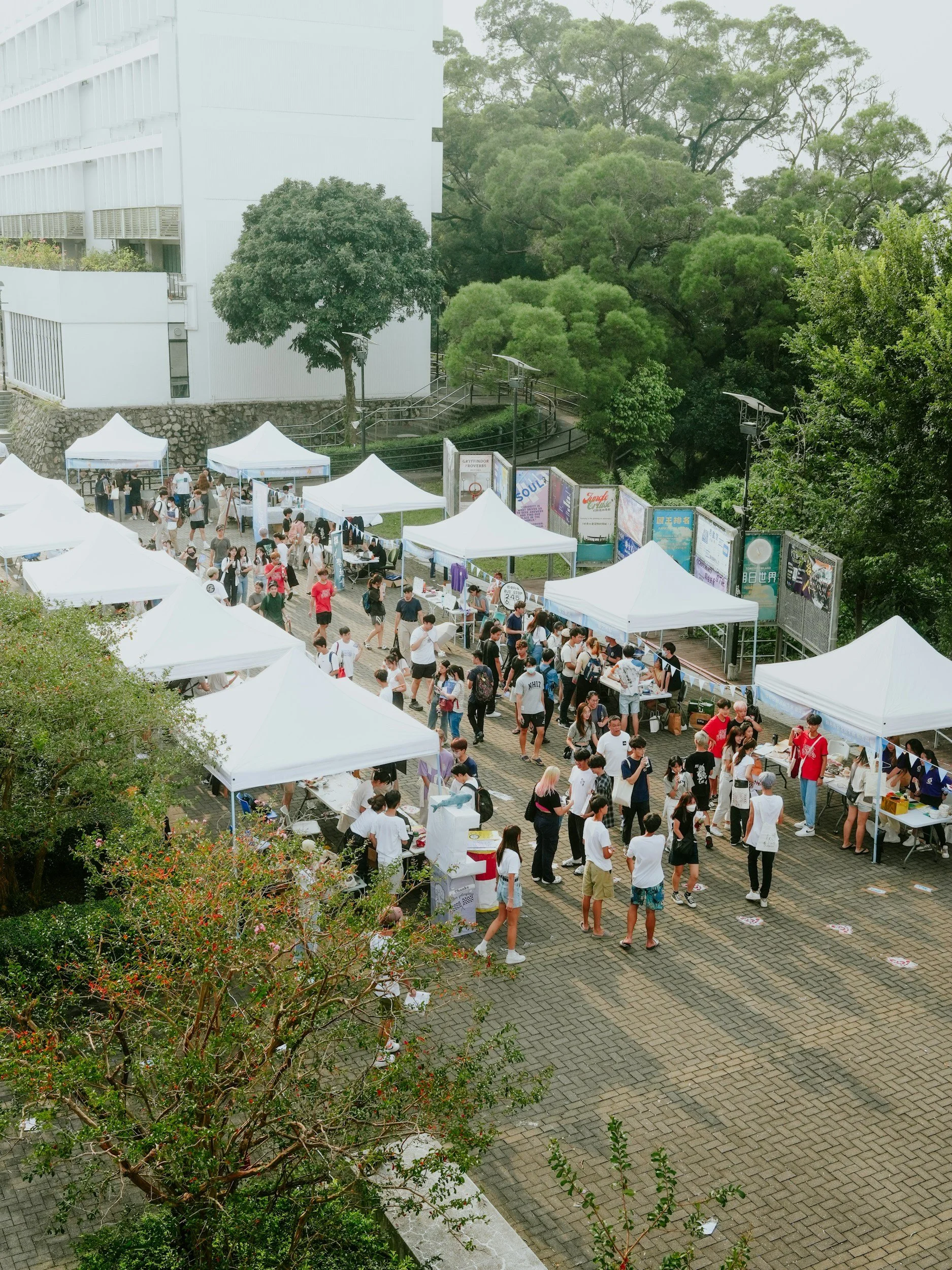 Outdoor event with multiple white tents and a crowd of people walking and browsing at stalls, set against a backdrop of green trees and a modern building.