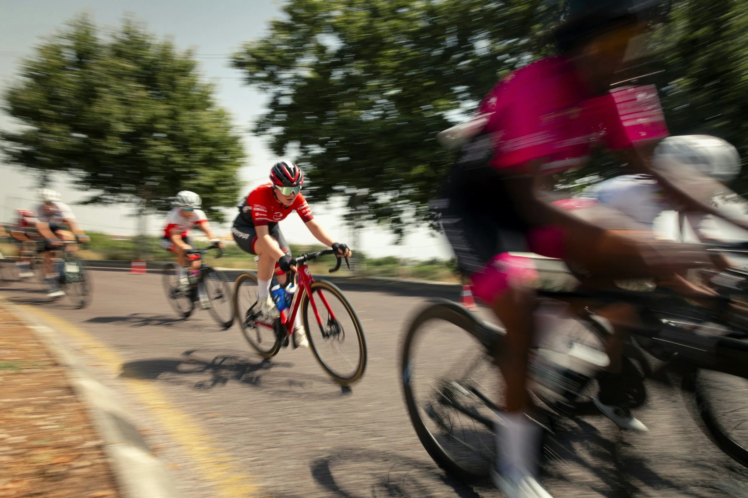 A group of cyclists riding on a road, some wearing helmets and sports gear. The image is blurred, indicating fast movement or motion.