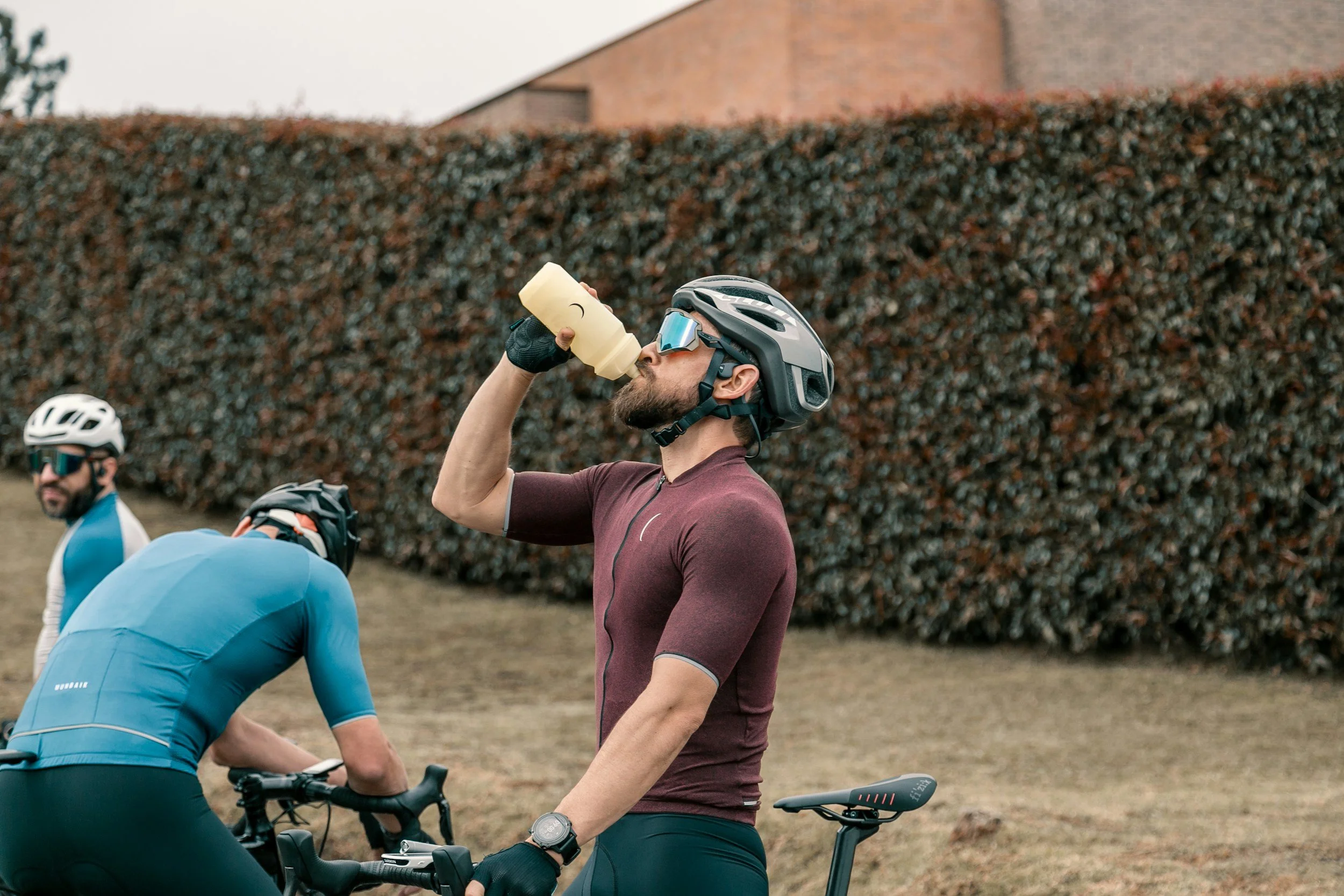 A man wearing a bike helmet and sunglasses drinking water from a bottle while taking a break during a cycling session. In the background, there are two other cyclists and a large hedge.