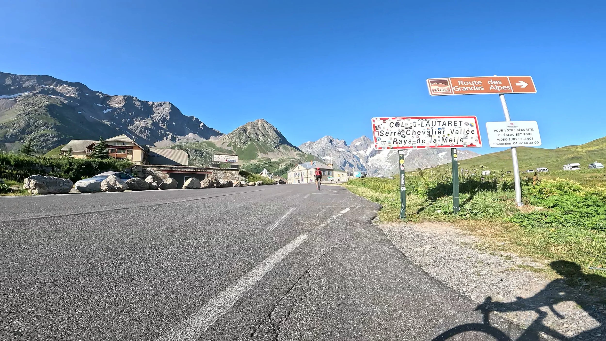 Mountain landscape with a clear blue sky, a winding mountain road, a cyclist riding, and signs indicating the route of the Great Alps.