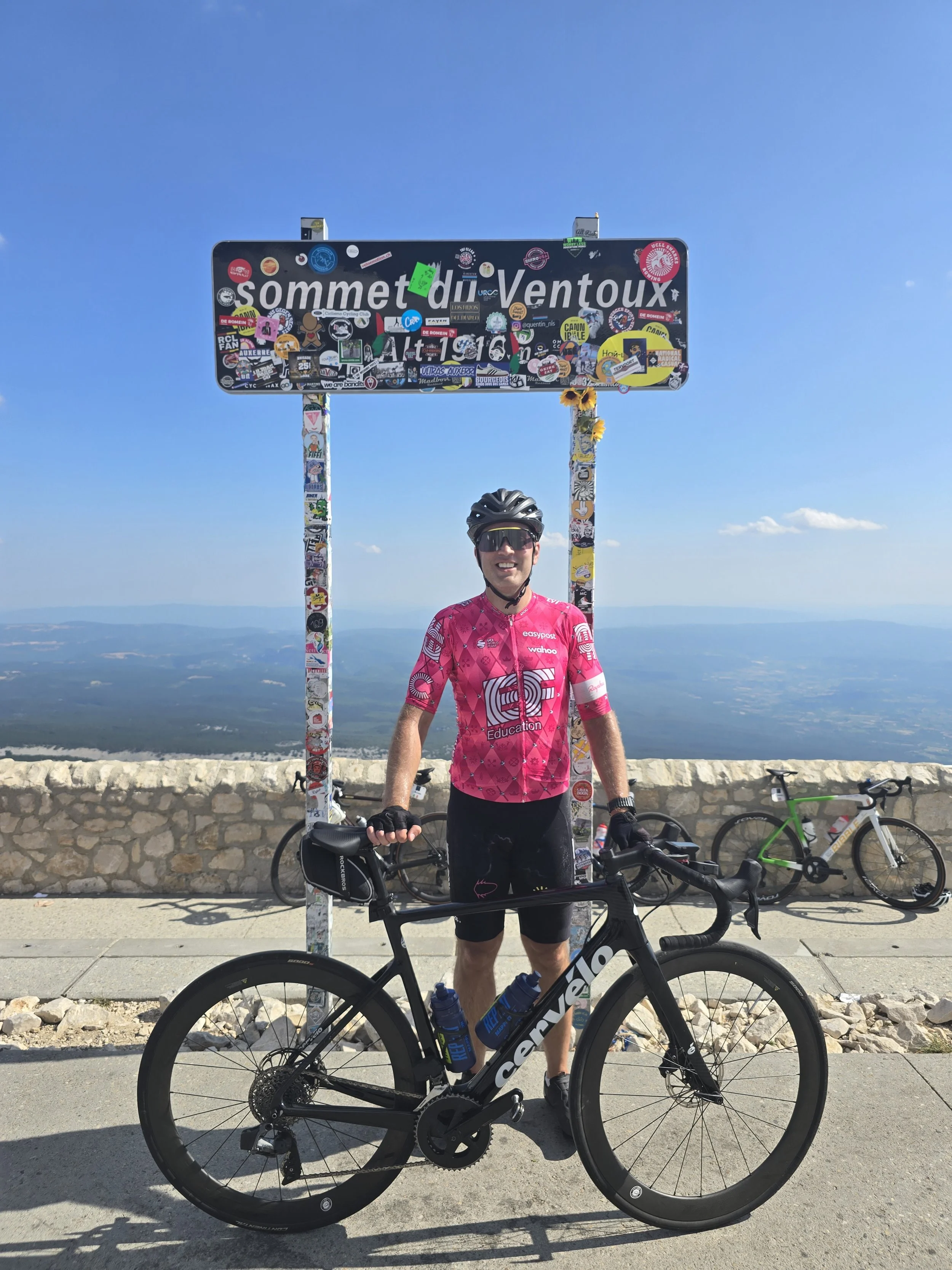 A man dressed in cycling gear, wearing a pink jersey and black shorts, stands next to a black road bike with drop handlebars and water bottles, on a mountain overlook. Behind him, a woman in a purple jersey and helmet, and other bicycles are visible. There is a signPost with a multitude of stickers and a clear sky in the background.