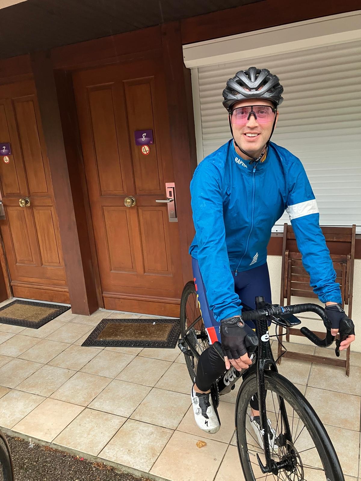 A man in a blue cycling jacket, black helmet, and pink sunglasses standing with a black racing bicycle indoors, with wooden doors and tiled floor in the background.