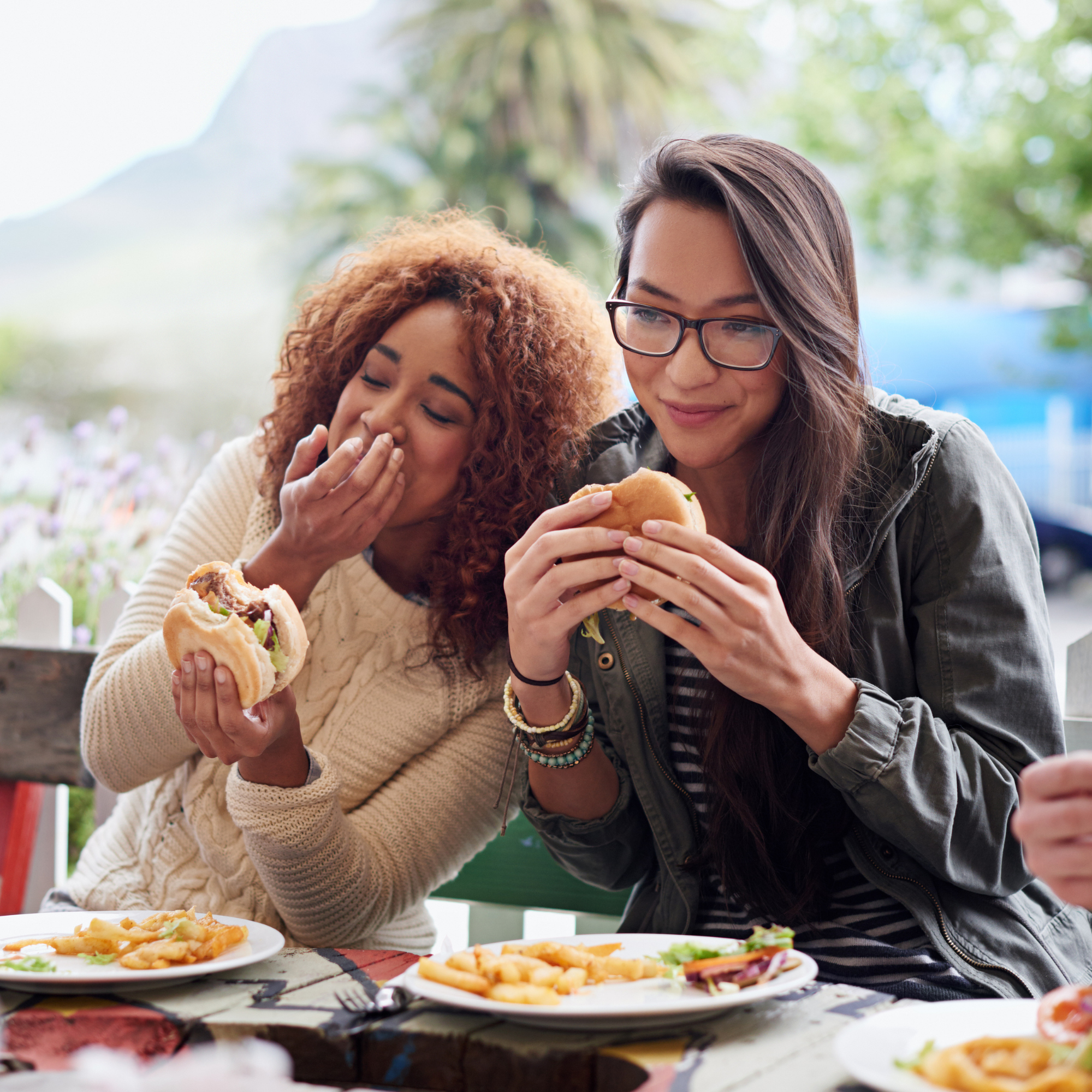 Two women sitting at an outdoor table, enjoying a meal with plates of food, surrounded by trees and bright natural light.