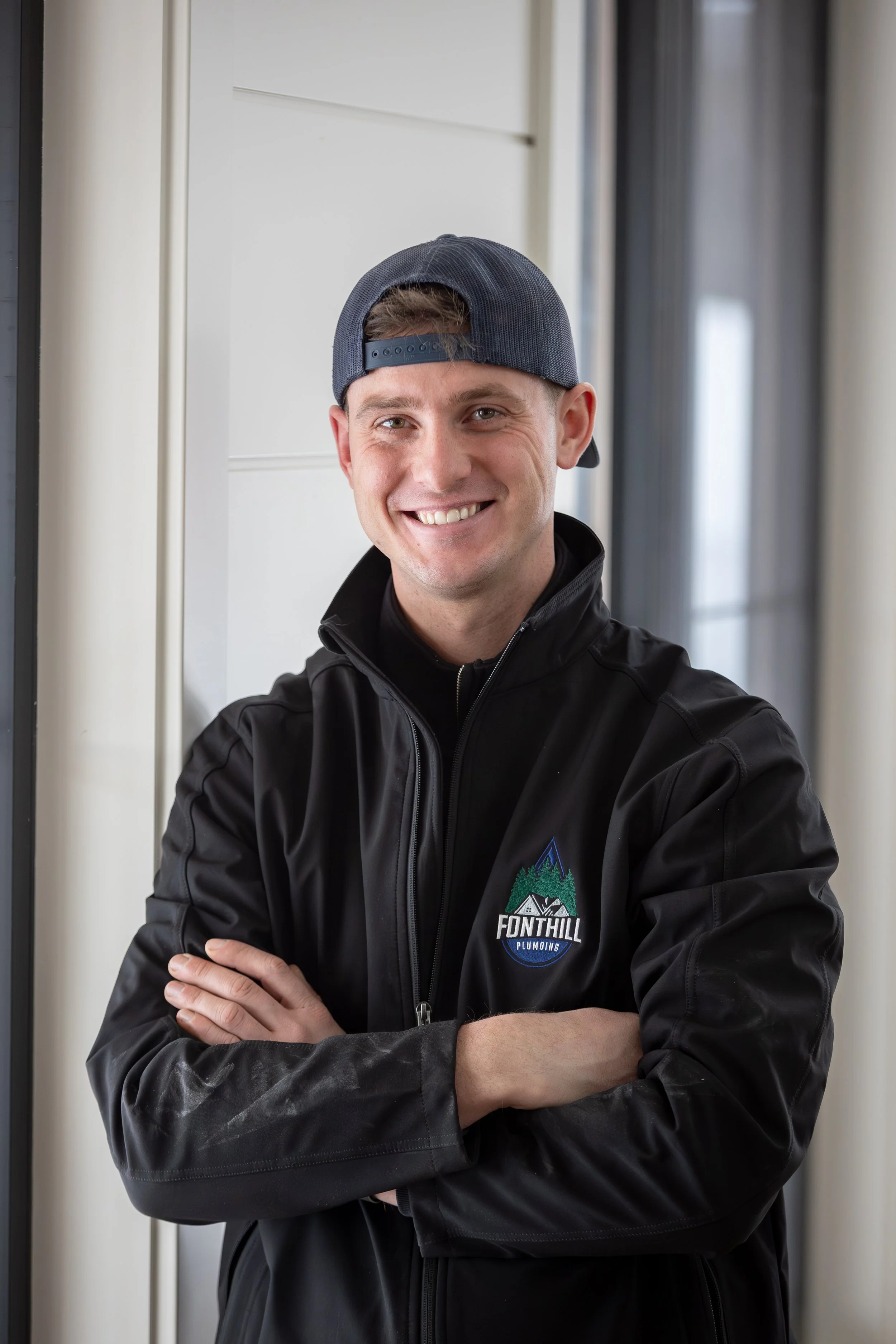 The owner/licensed plumber wearing a backward baseball cap and a black jacket with a 'Fonthill Plumbing' logo, standing indoors with arms crossed.