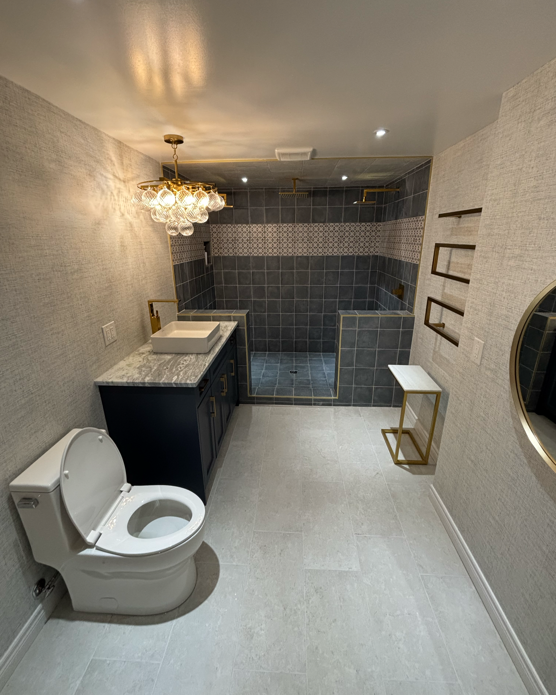 Modern bathroom with a toilet, marble countertop with sink, decorative tile wall, and a glass enclosed shower area with gold fixtures. There is a chandelier, wall sconce, and floating shelves.