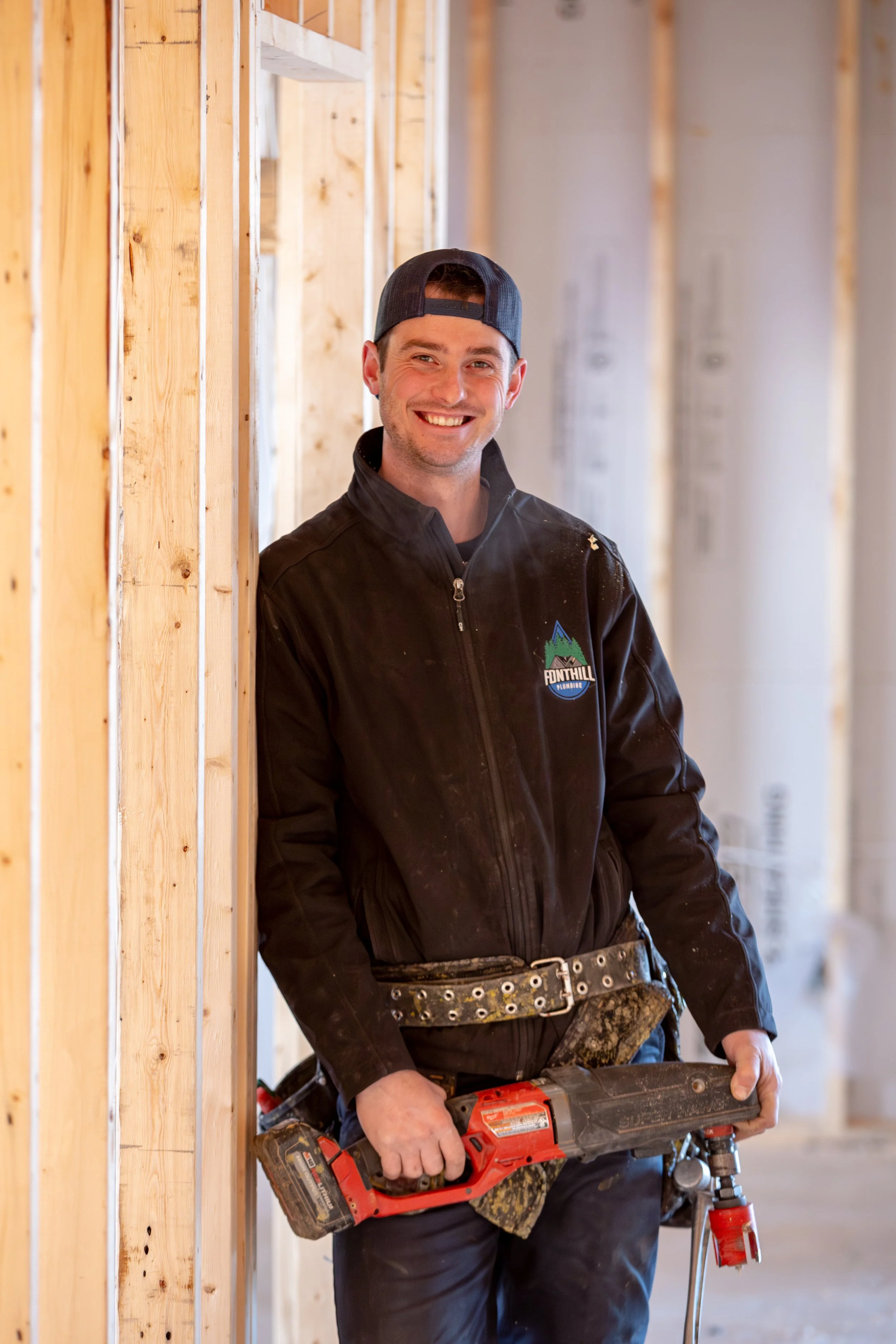 The owner/licensed plumber is wearing a black jacket with the Fonthill Plumbing logo on the front. He is wearing a snapback and smiling while standing among wooden framing at a construction site, holding a power tool.