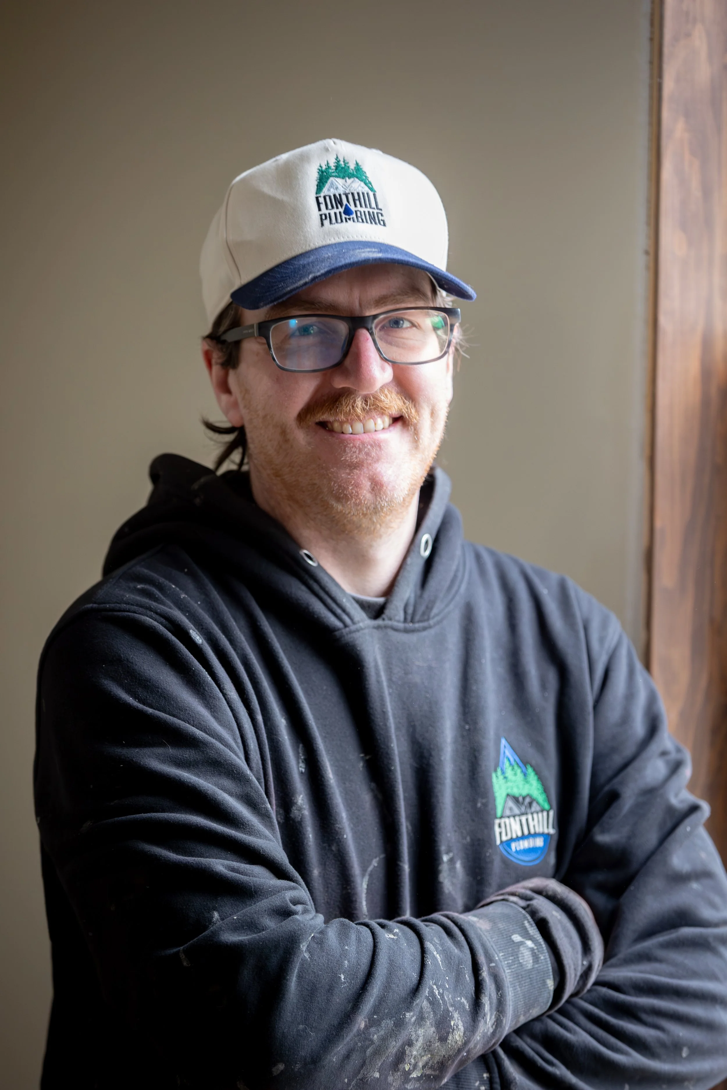 A plumber with glasses and a beard smiling, wearing a white trucker hat with a logo reading 'Fonthill Plumbing' and a black hoodie with the same logo, sitting indoors near a wooden wall.