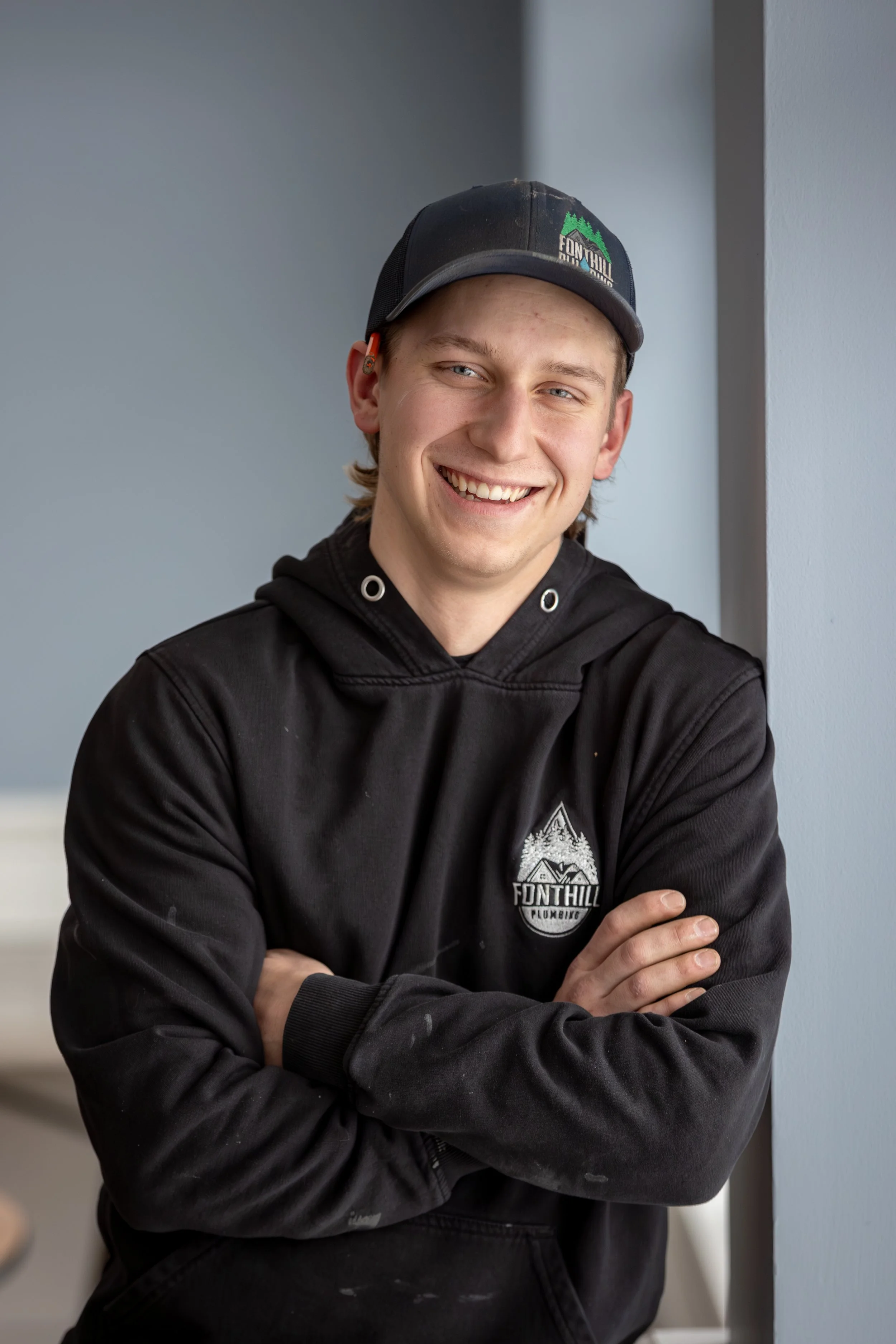 A smiling young plumber with light hair, wearing a black hoodie with a 'Fonthill Plumbing' logo, and a black cap, standing with his arms crossed against a plain light blue wall.