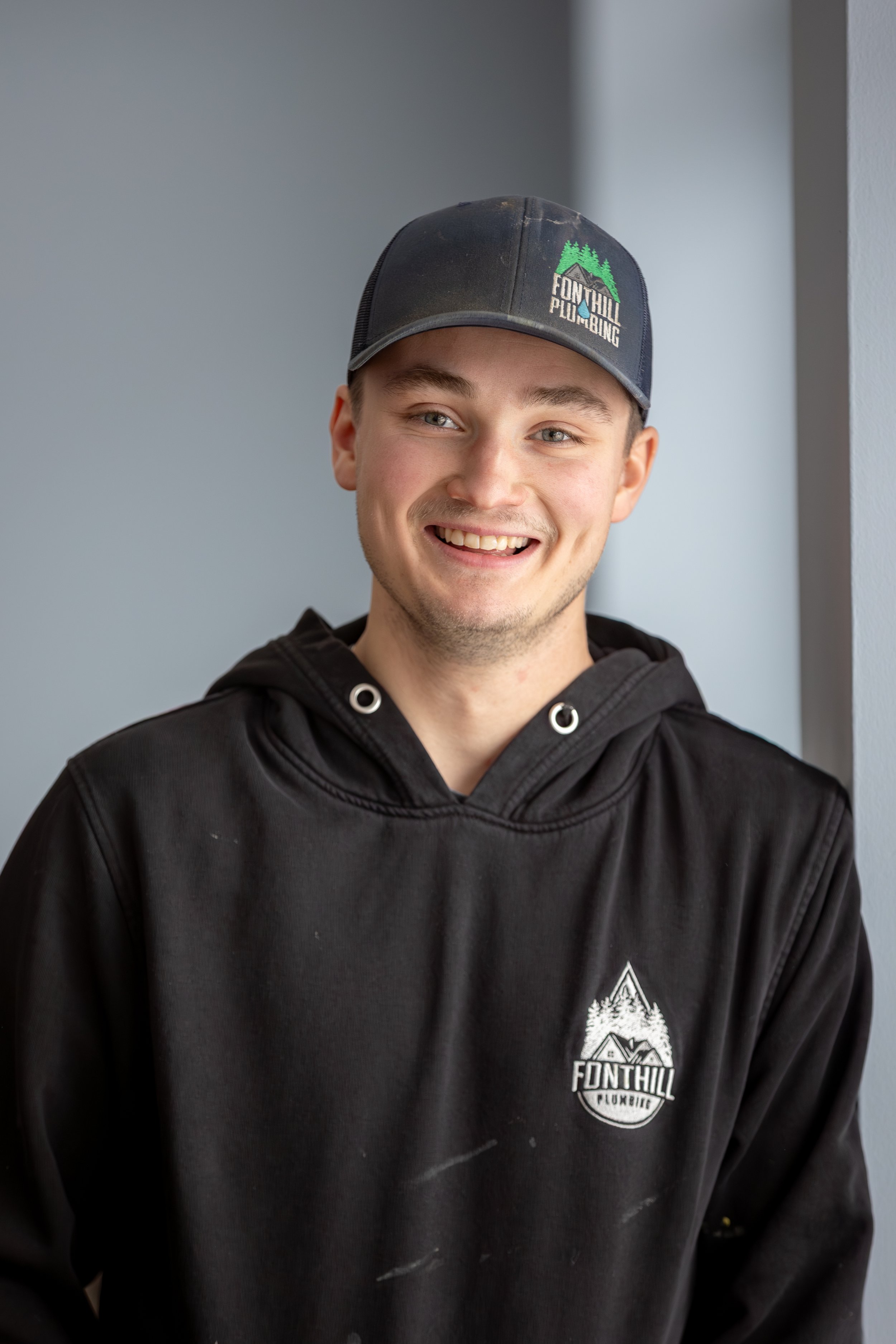 A young plumber wearing a black hoodie and a black baseball cap with a logo that reads 'Fonthill Plumbing'. He is smiling and standing indoors near a light gray wall.