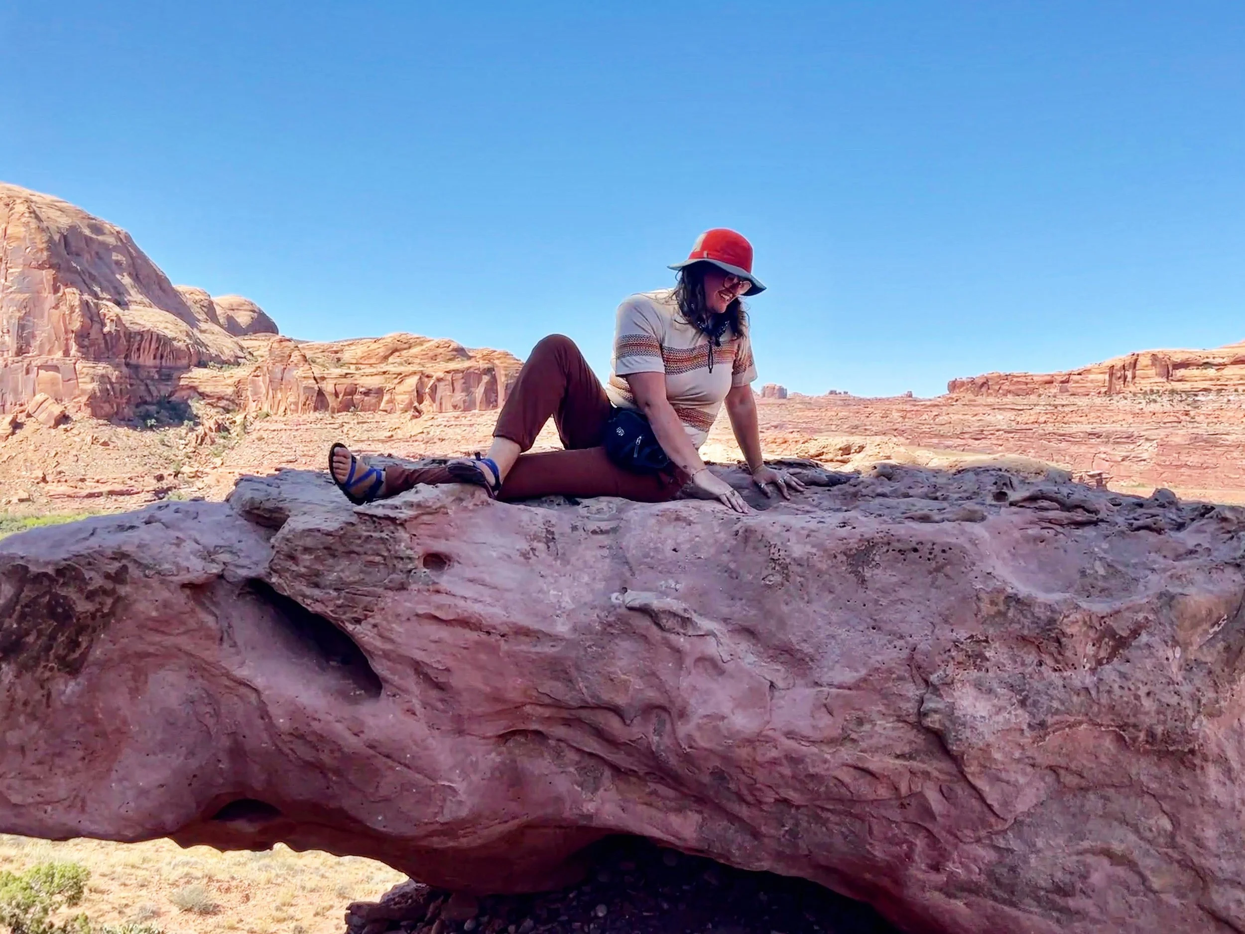 A woman in casual outdoor clothing, including a red hat, glasses, and brown pants, stretching on a large reddish rock formation in a desert landscape with red cliffs and a clear blue sky.