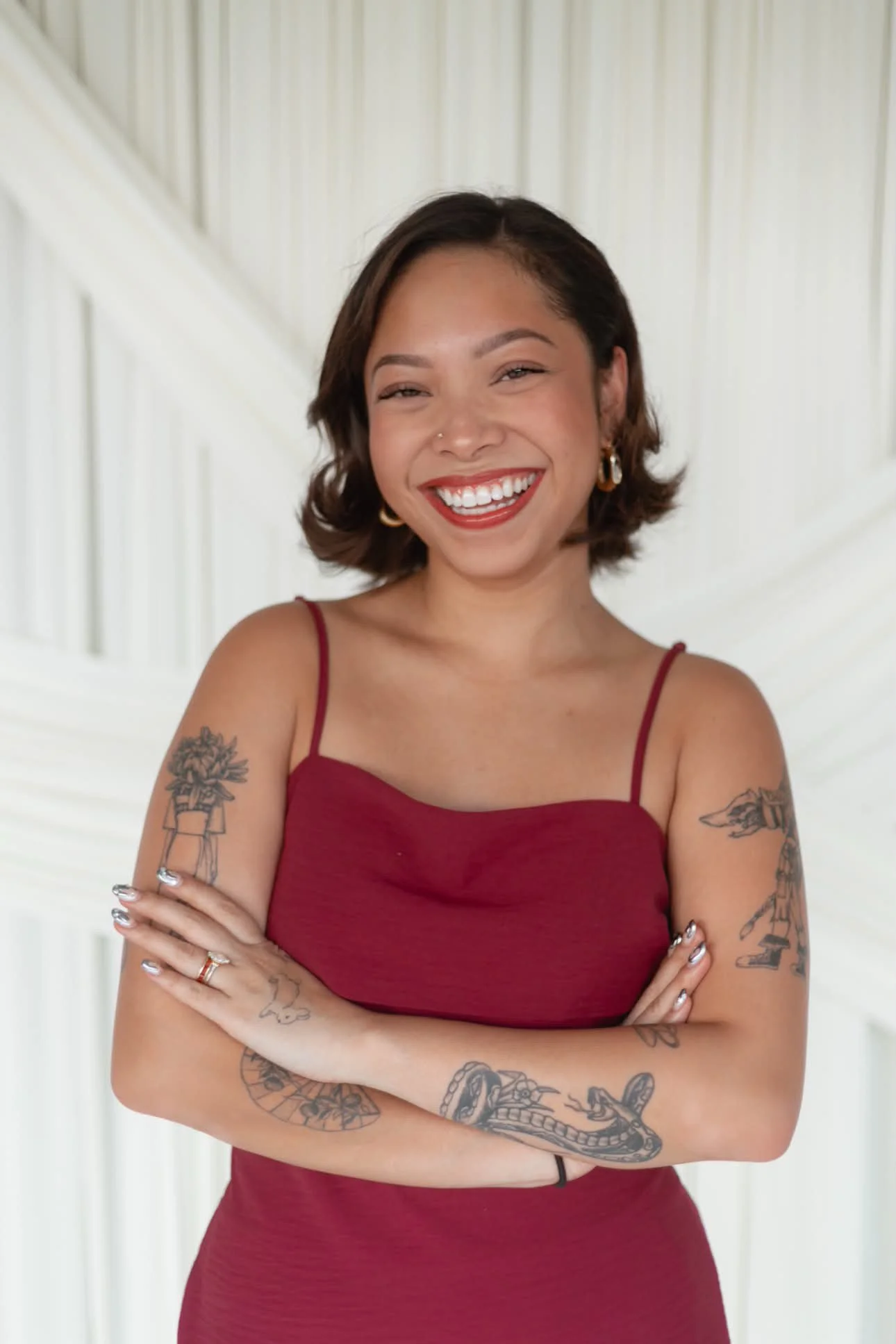A smiling woman with tattoos on her arms, wearing a red spaghetti strap top, standing indoors in front of a white wall with decorative molding. Aika Luxe Co Branding headshot