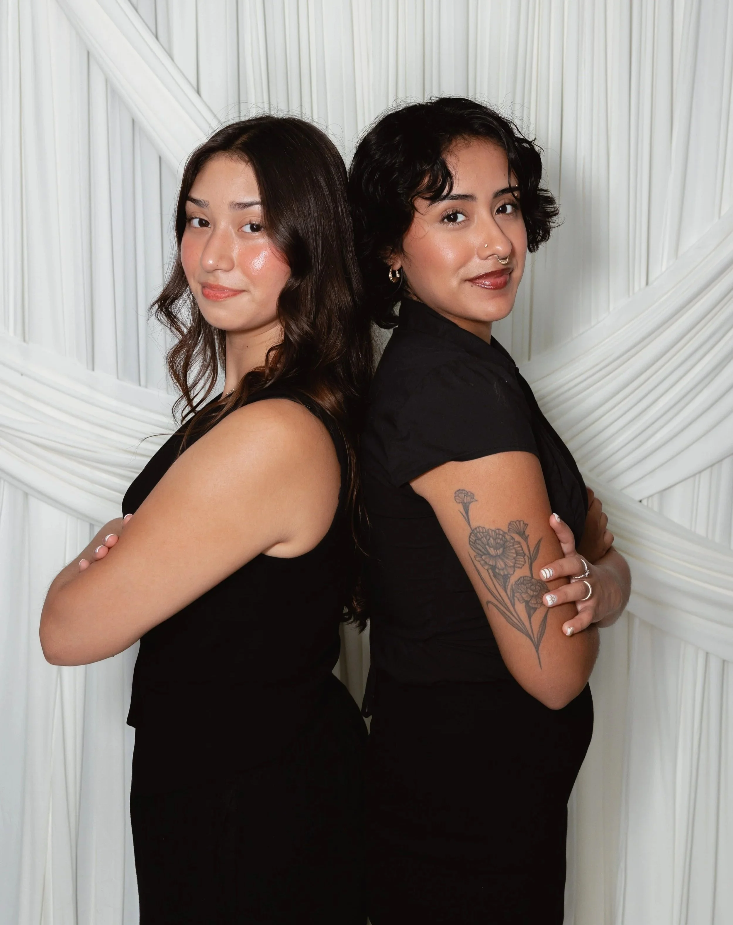 Two women standing back to back with arms crossed, smiling slightly, against a white textured background.