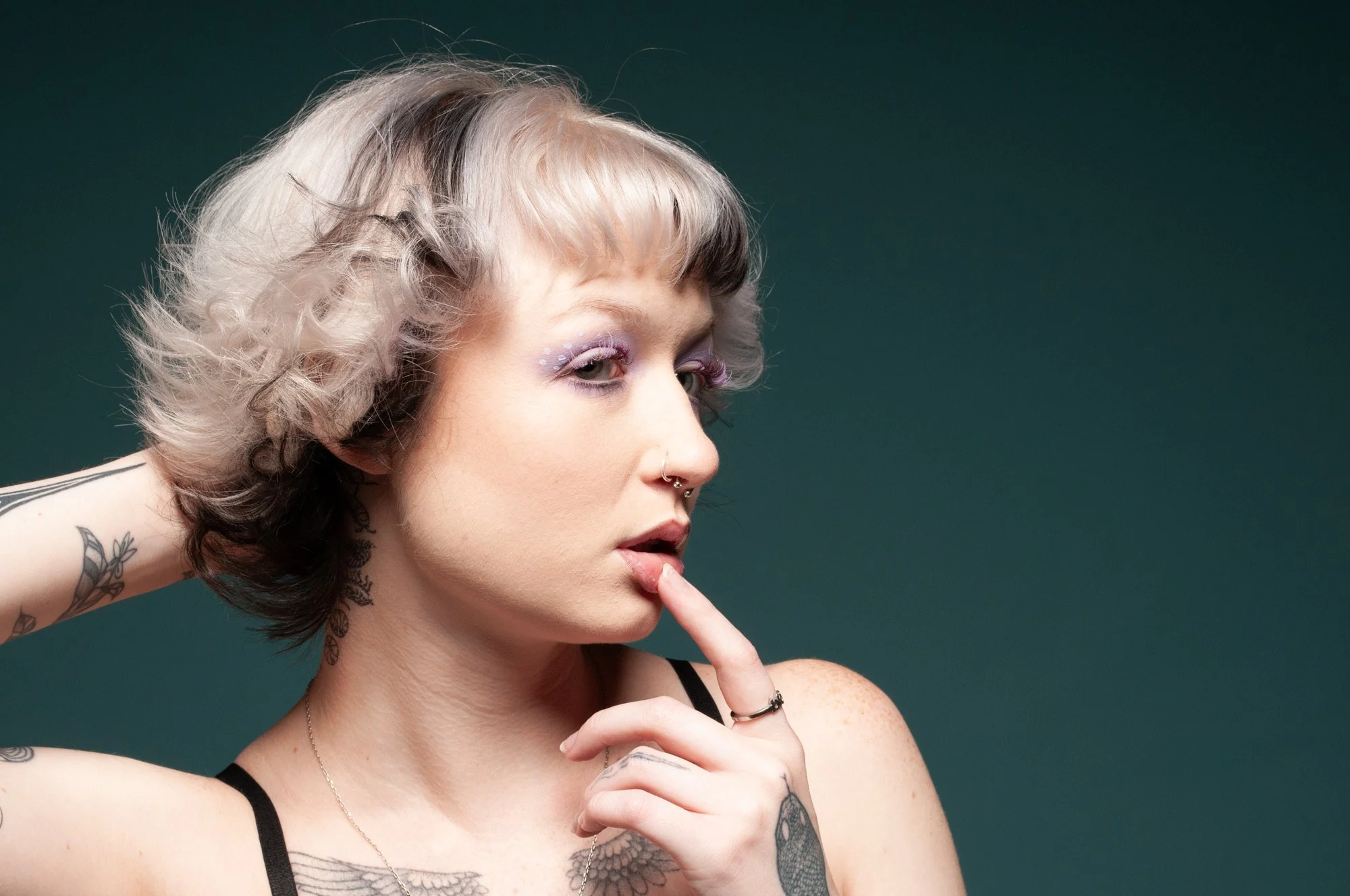 A woman with short, curly, two-toned hair (blonde and black) is posing with her finger on her lips, against a dark green background. She has tattoos on her shoulder and neck, eye lash extensions by Ica ( Aika Luxe Co ) in Bentonville