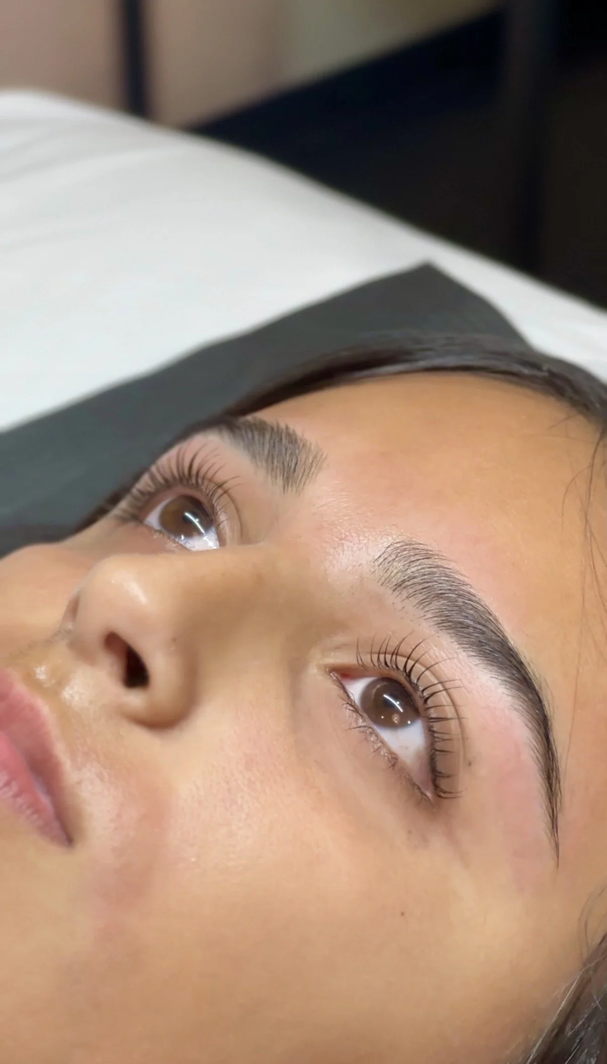 Close-up of a woman's face lying down on a pillow, showing her brown eyes, well-groomed eyebrows, and clear skin. Brow shaping and lash lift.