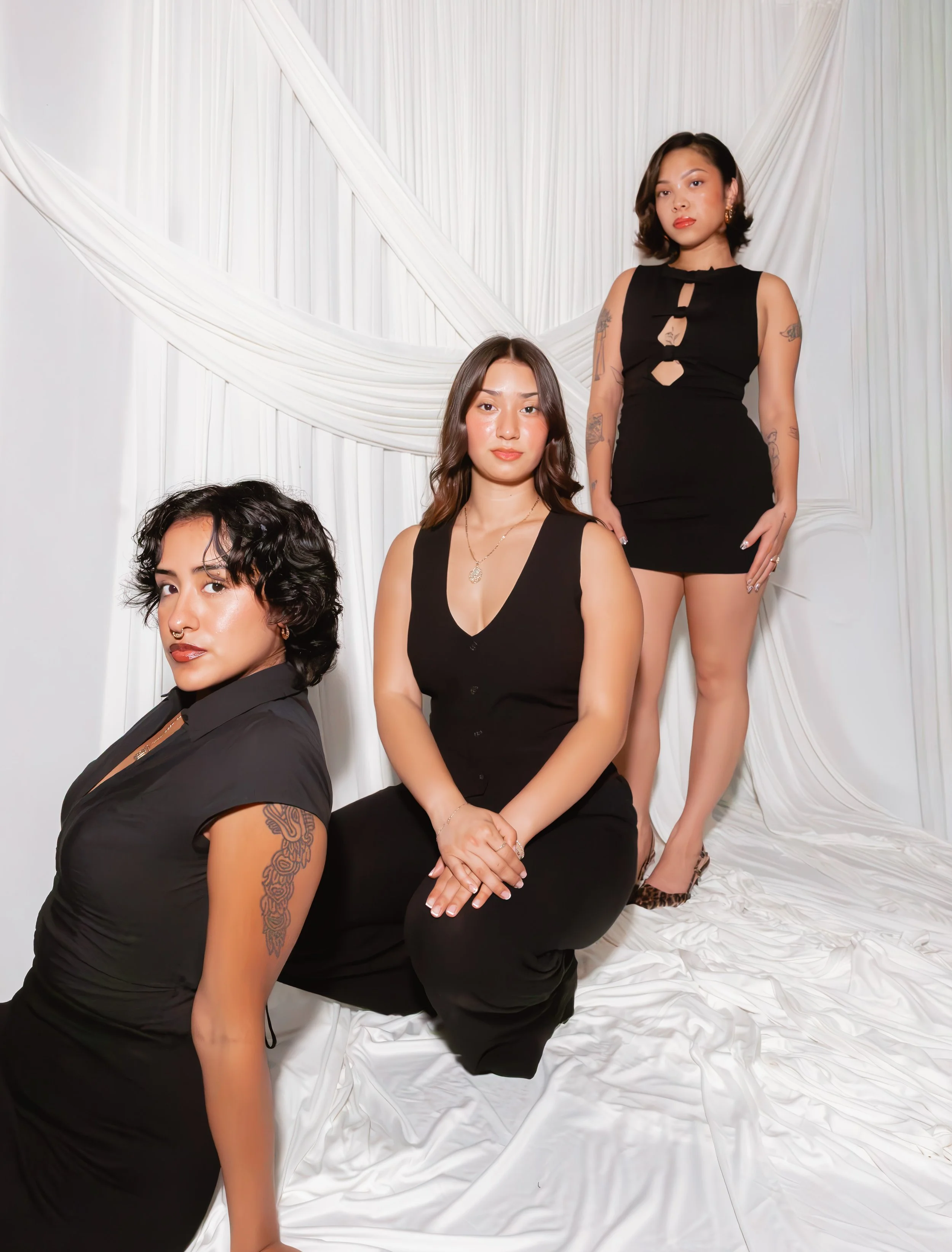 Three women in black dresses posing against a white fabric backdrop. Aika Luxe Co Team Members