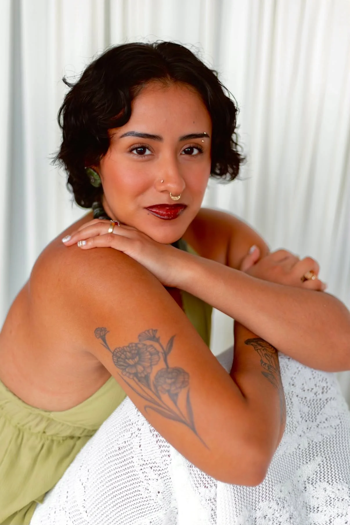 A woman with short dark curly hair, tattoos on her arm, and dark lipstick, resting her arms on a white table, looking at the camera, with a cream-colored background. Aika Luxe Co Branding headshot