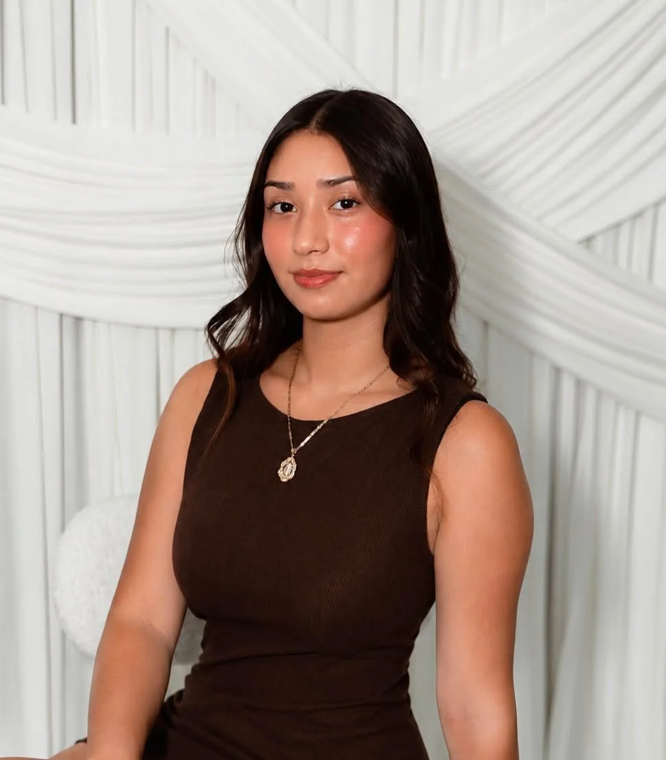 A woman with shoulder-length dark hair and a light skin tone, wearing a sleeveless brown dress and a necklace, sitting against a white textured background. Aika Luxe Co Branding headshot
