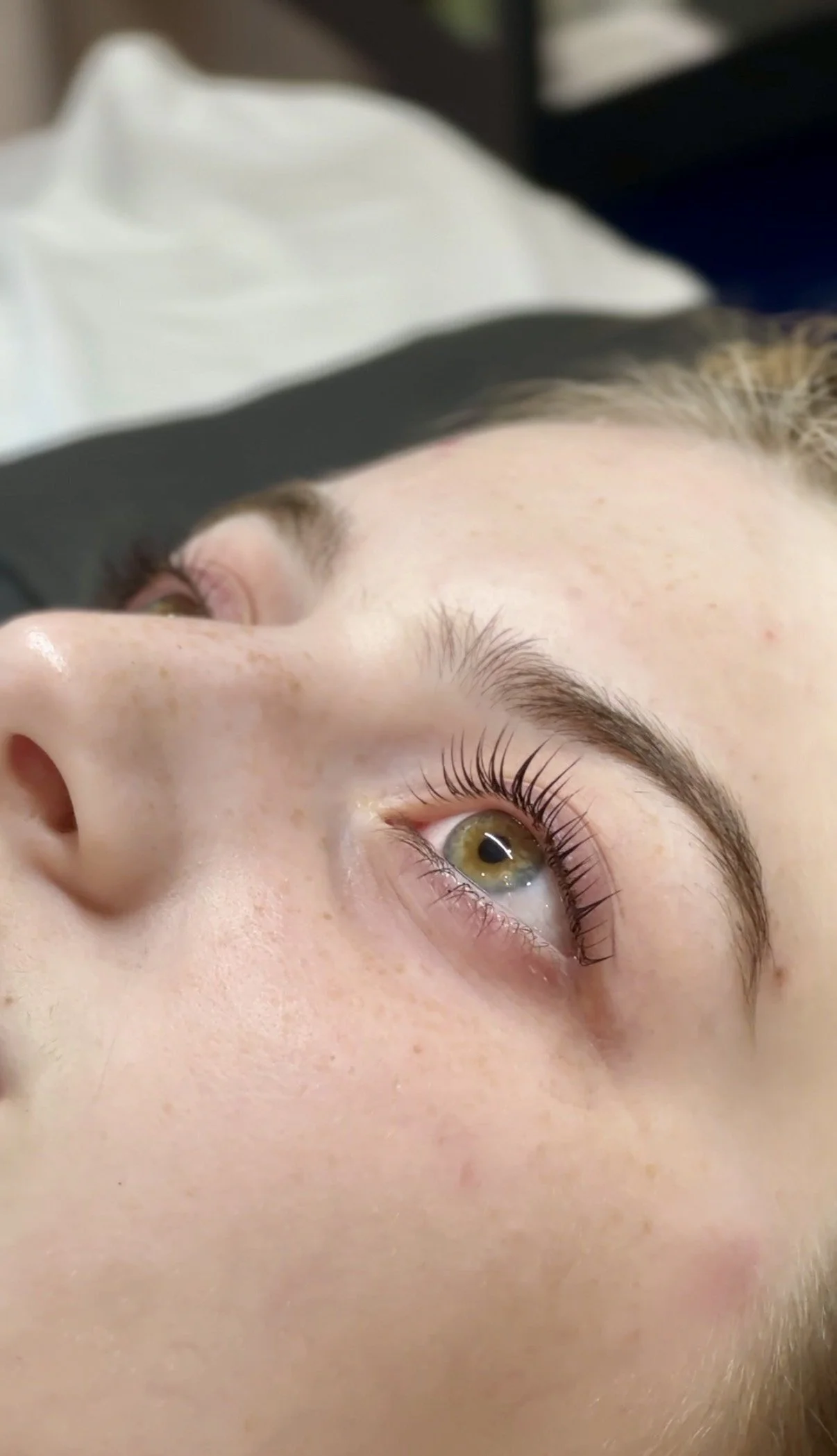 A close-up of a person's face showing one eye with long eyelashes, showing a greenish eye color, and clear skin with freckles, lying on a surface with a white and black cloth nearby. Brow shaping and lash lfit.