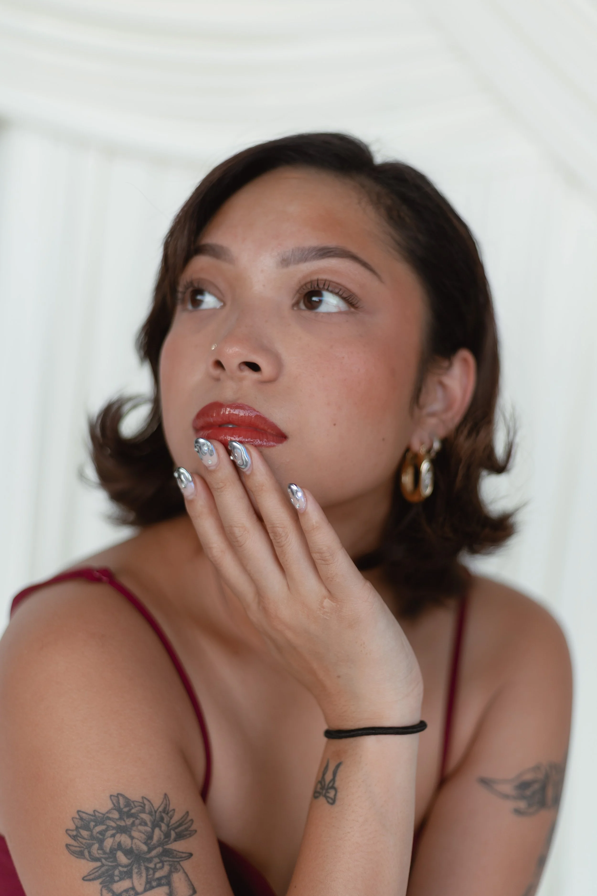 A young woman with short, dark brown hair styled in soft waves, wearing a red spaghetti strap top. She has tattoos on her arms, including a large flower on her left arm and a bird on her right forearm. She has a light skin tone, makeup with red lipstick, and metallic nail polish. She is touching her chin with her left hand, looking upwards and to her right against a light, plain background.
