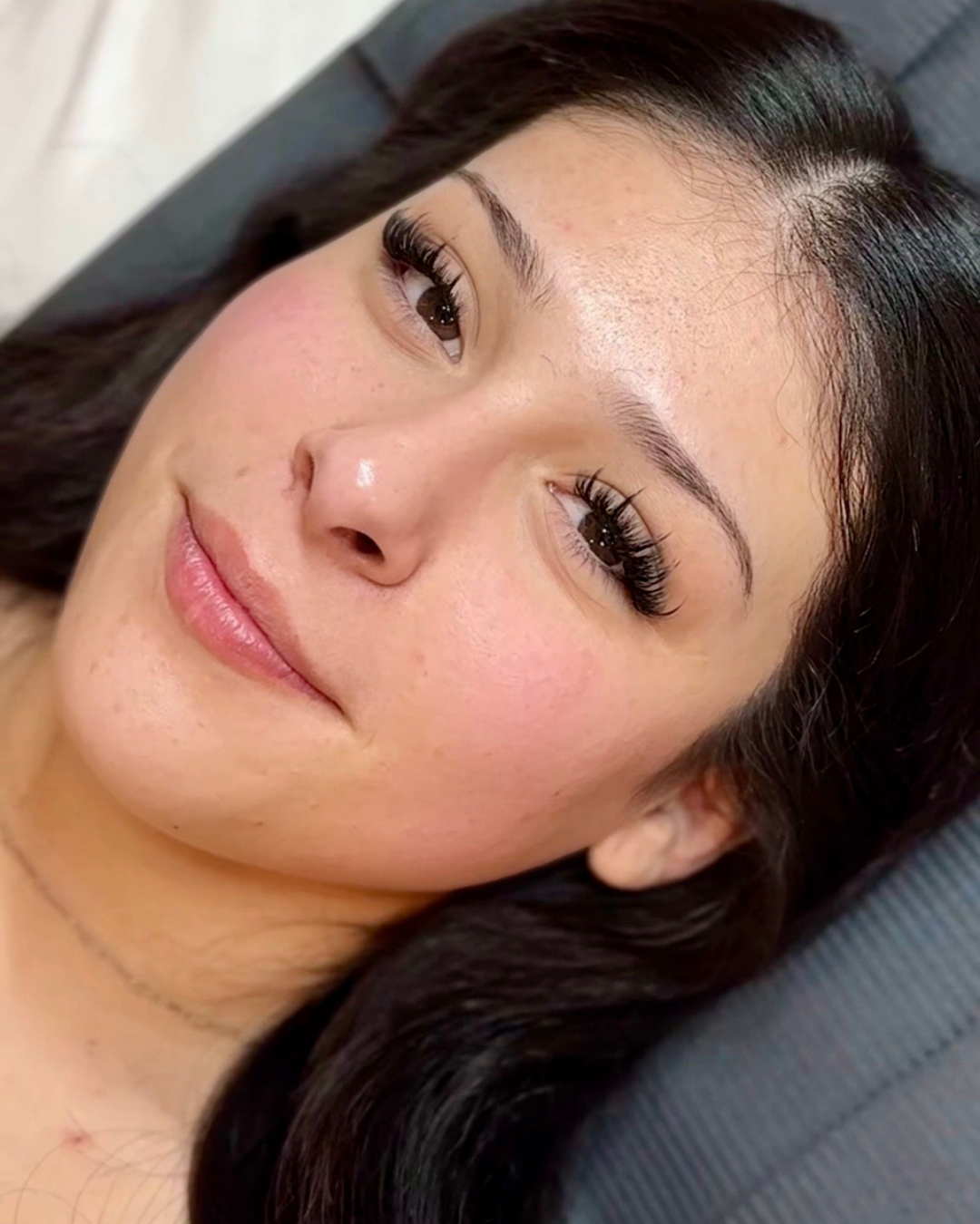 A close-up of a woman with long, dark hair, clear skin, and well-groomed eyebrows, lying on a gray surface, smiling softly.