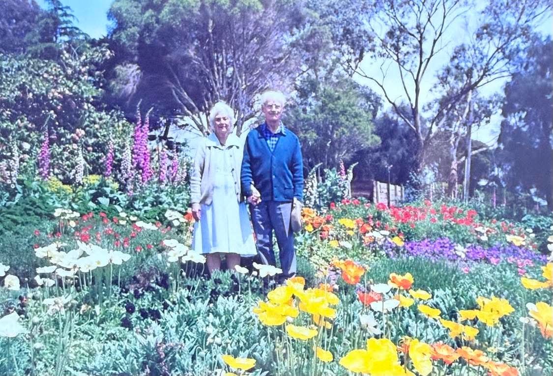 An elderly couple standing in a vibrant garden with various colorful flowers and trees in the background.
