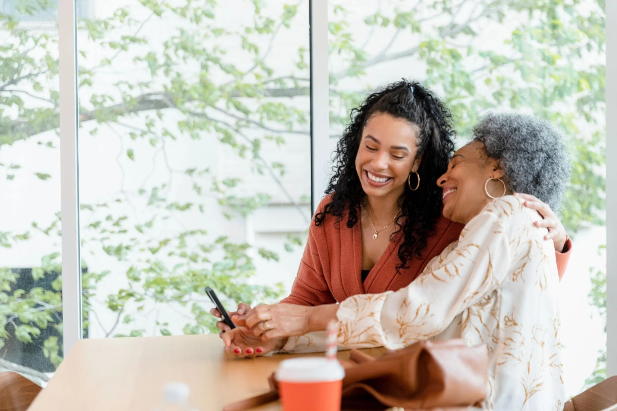Two women, one younger with dark curly hair and hoop earrings, and one older with gray curly hair and hoop earrings, are sitting at a table in front of a large window with green trees outside. They are smiling and hugging each other, with the younger woman holding a smartphone.