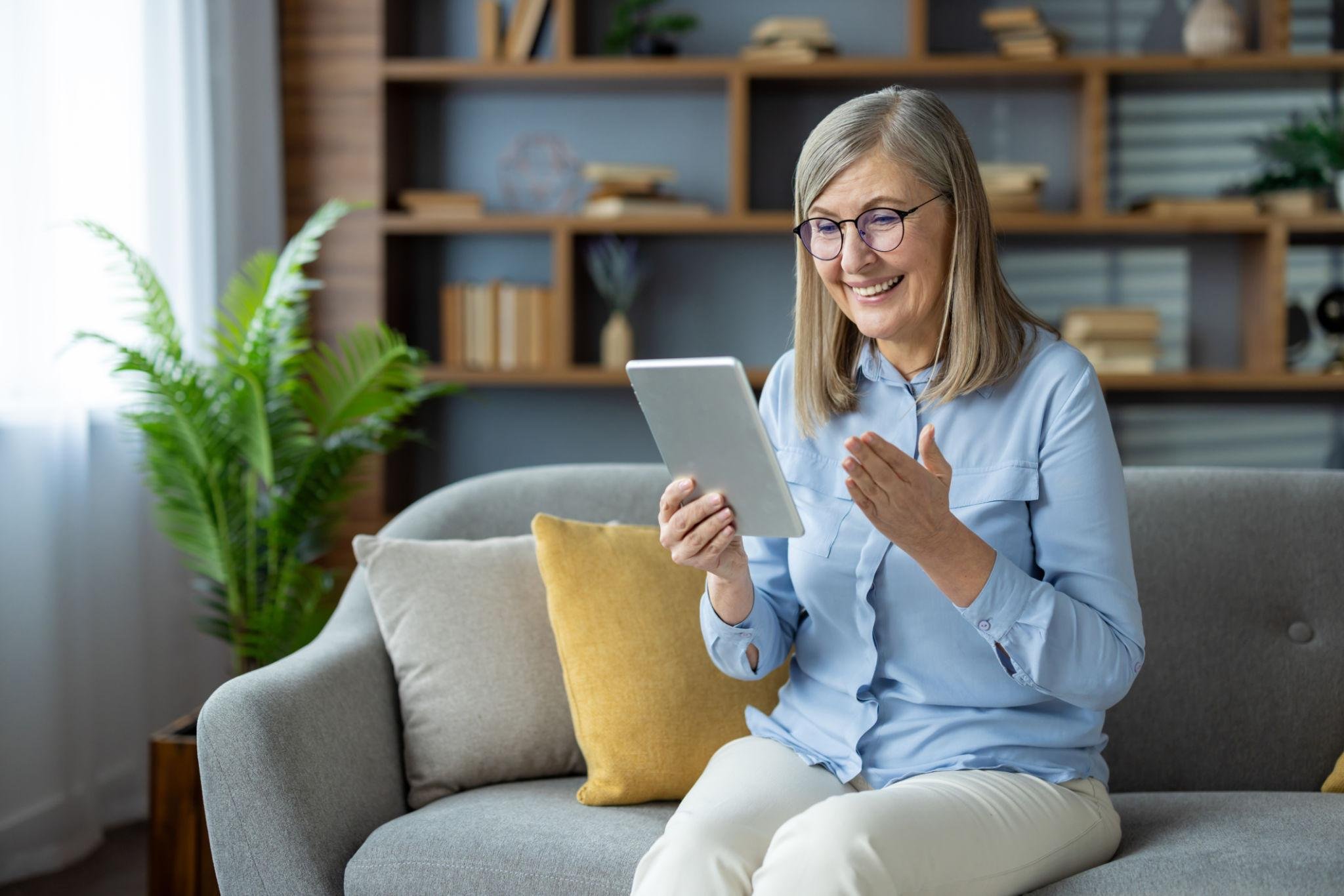 A smiling woman with gray hair and glasses sitting on a gray couch, looking at a tablet in her hand, in a cozy living room with wooden shelves, books, and plants in the background.