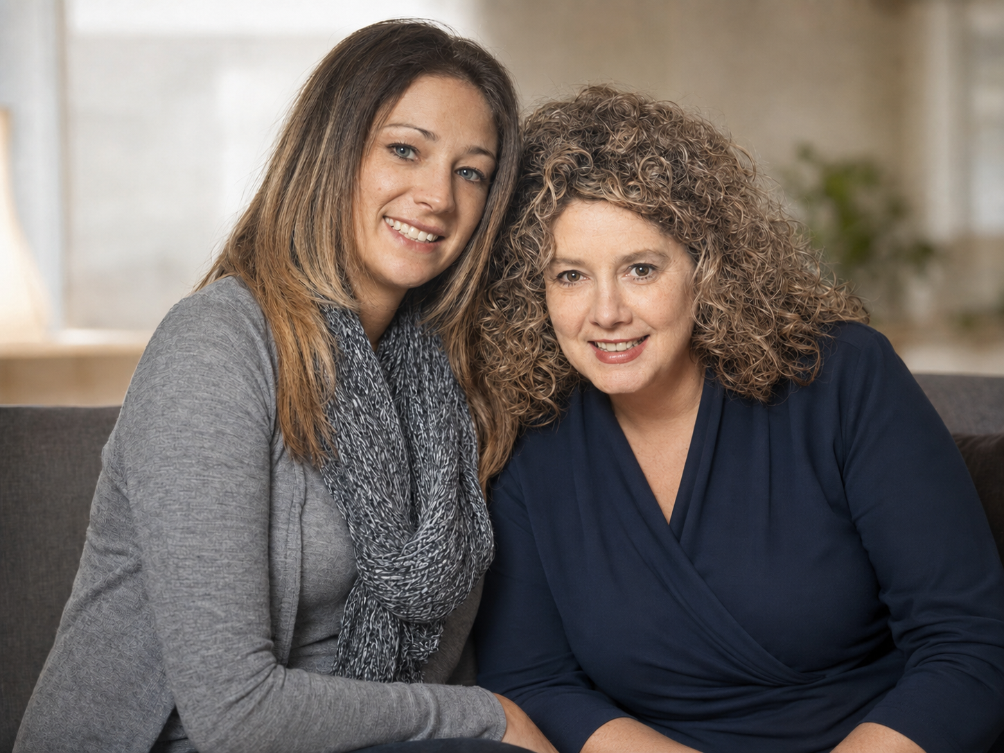 Two women sitting close together on a sofa, smiling at the camera in a well-lit indoor setting.