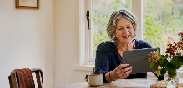 Older woman sitting at a dining table, looking at a tablet, with a mug and a vase of flowers on the table, and a window with greenery outside.