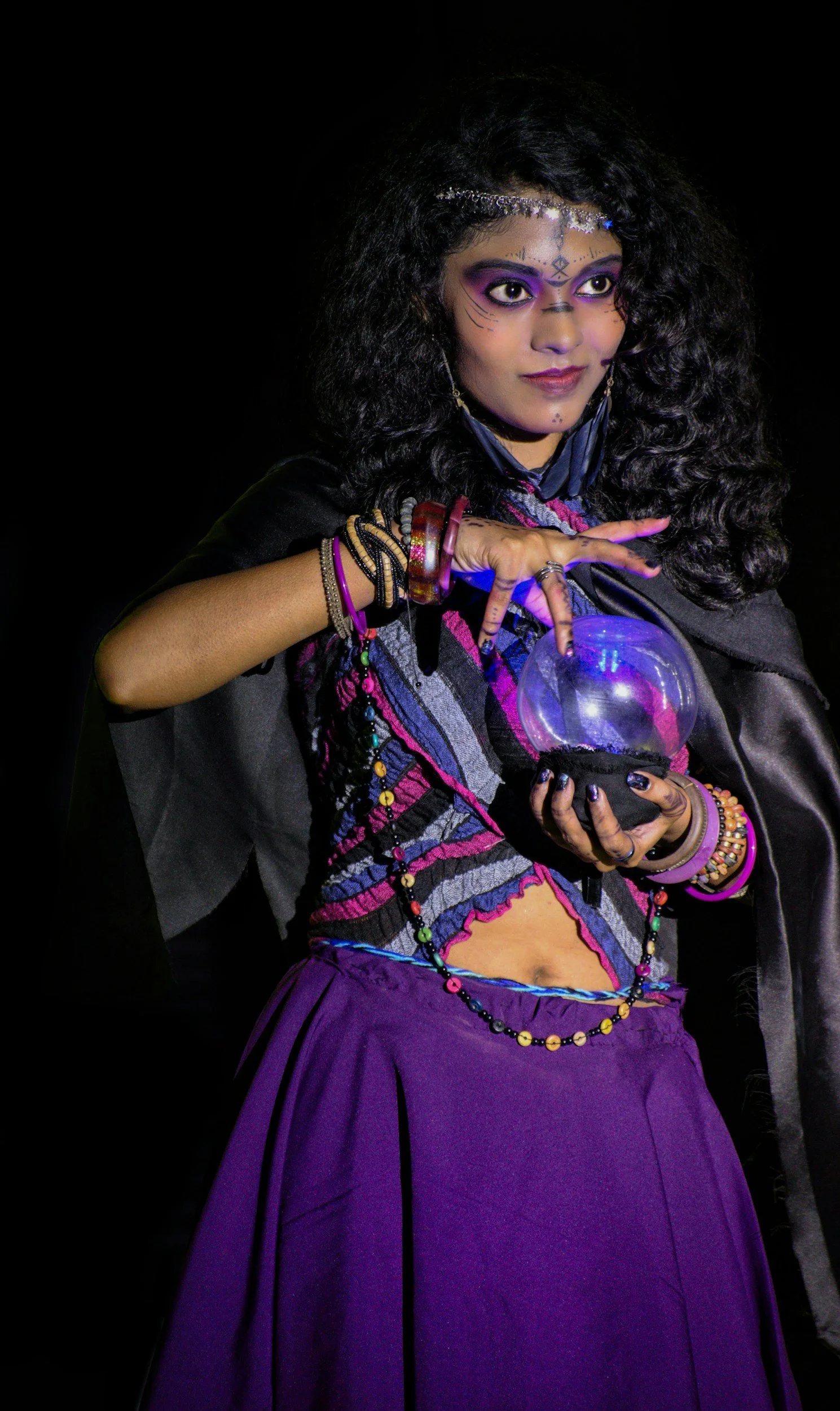 Woman dressed as a fortune teller with colorful makeup, layered jewelry and a crystal ball against a dark background.