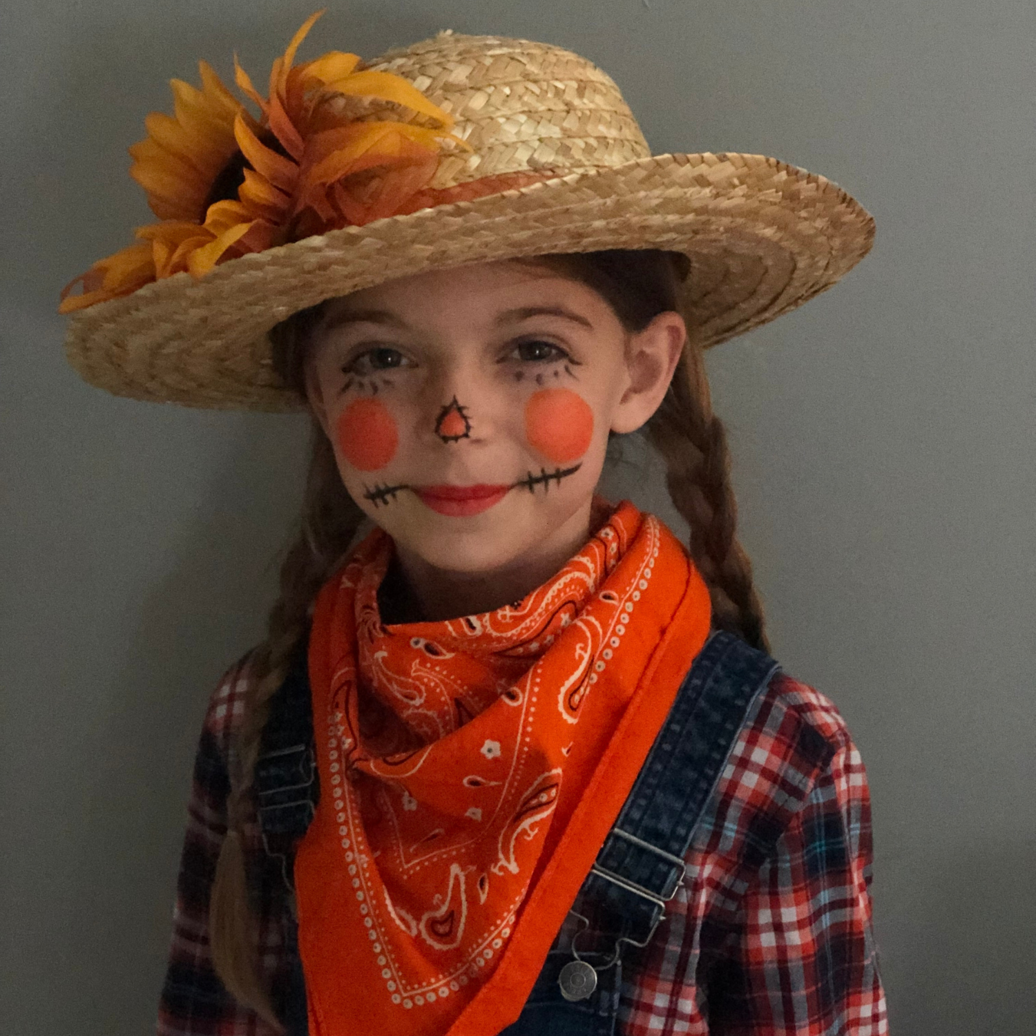 Girl dressed as a scarecrow in overalls, a plaid shirt and straw hat,  standing in front of a gray wall.