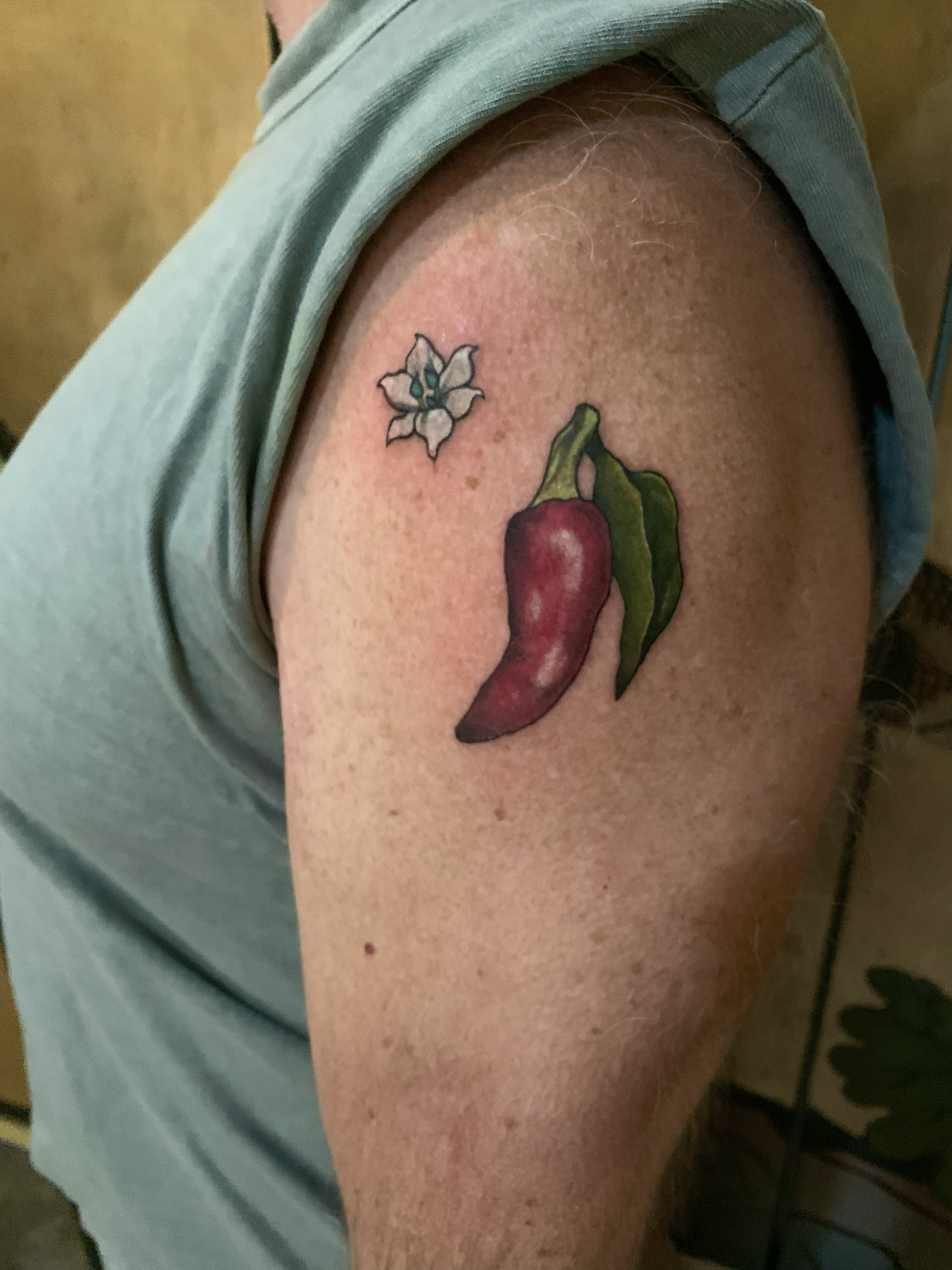 Small red chili pepper tattoo with a white flower on the upper arm