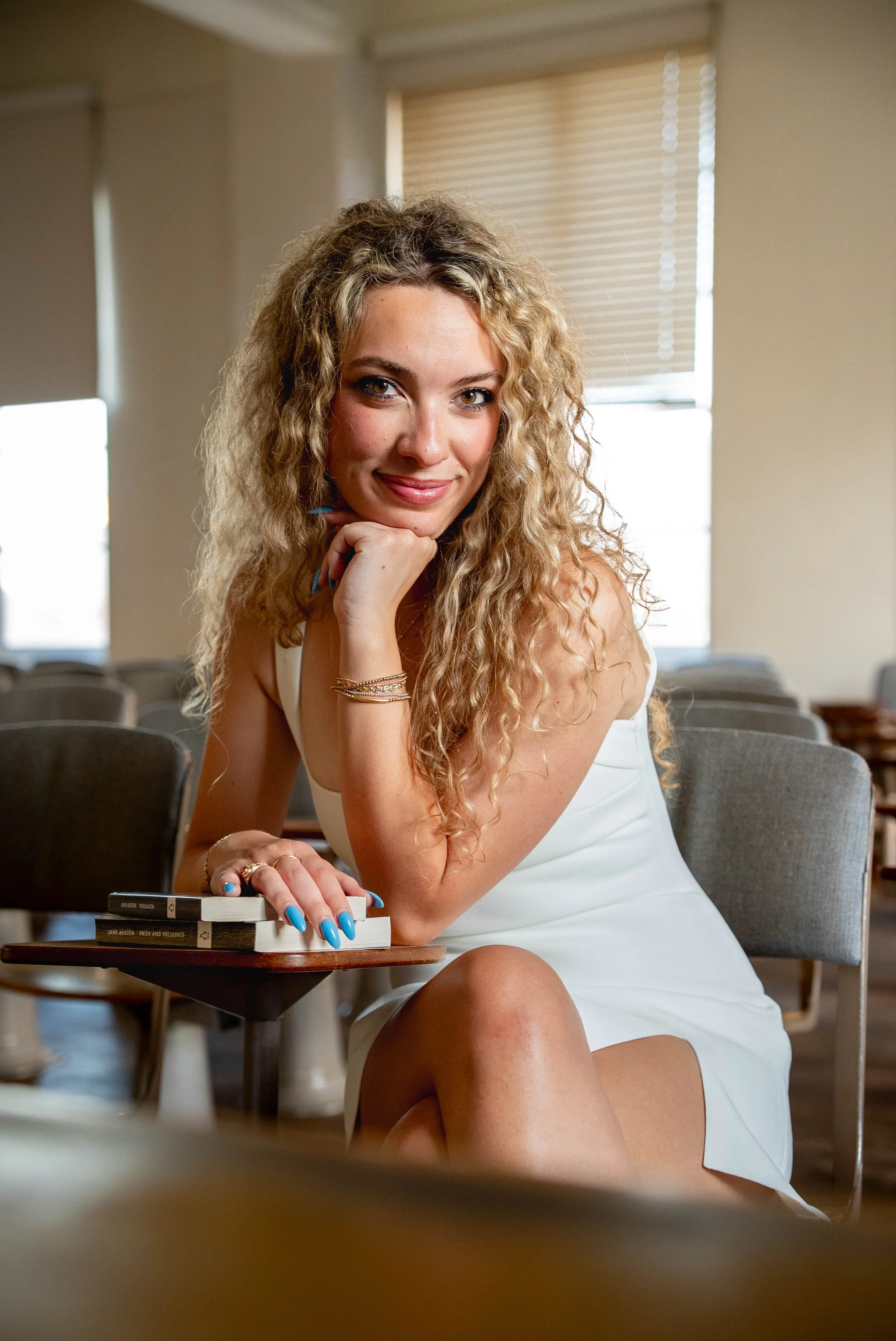 A woman with curly blonde hair, wearing a white dress, sitting at a table in a room with beige walls and window blinds, smiling and resting her chin on her hand.