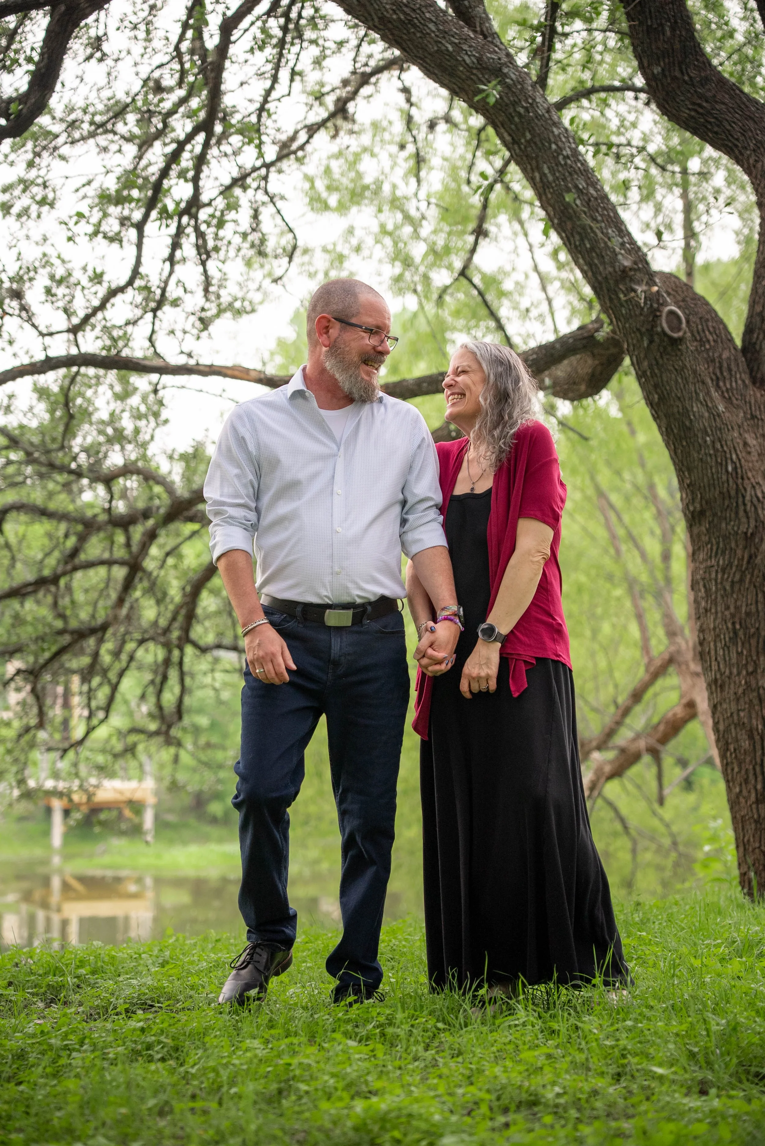 A smiling middle-aged man and woman holding hands outdoors under a large tree with green leaves, standing on grass near a small pond, enjoying a joyful moment together.