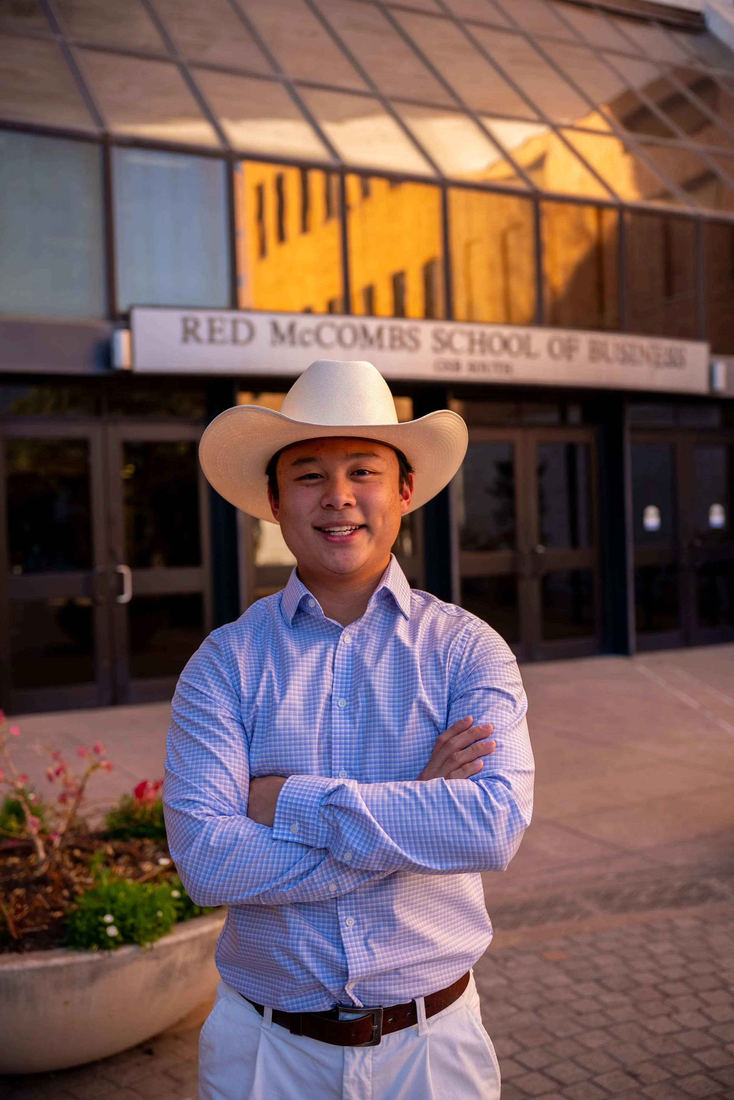 Young man wearing a white cowboy hat, a blue checkered shirt, and white pants with a brown belt standing outside in front of a building with a sign that reads 'Red McCombs School of Business.' He is smiling with his arms crossed.
