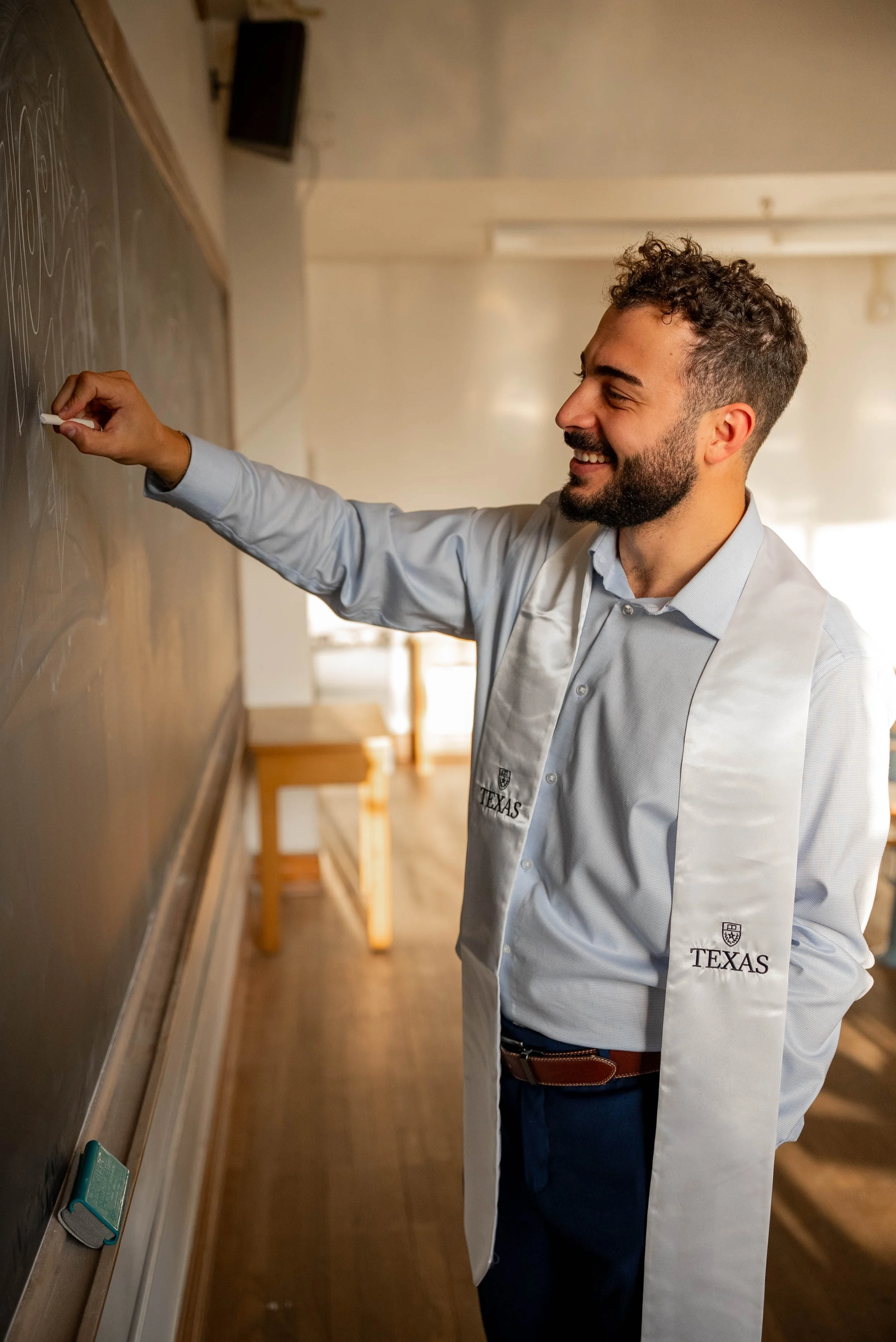 A man wearing a white lab coat with Texas insignia is smiling while writing on a blackboard.