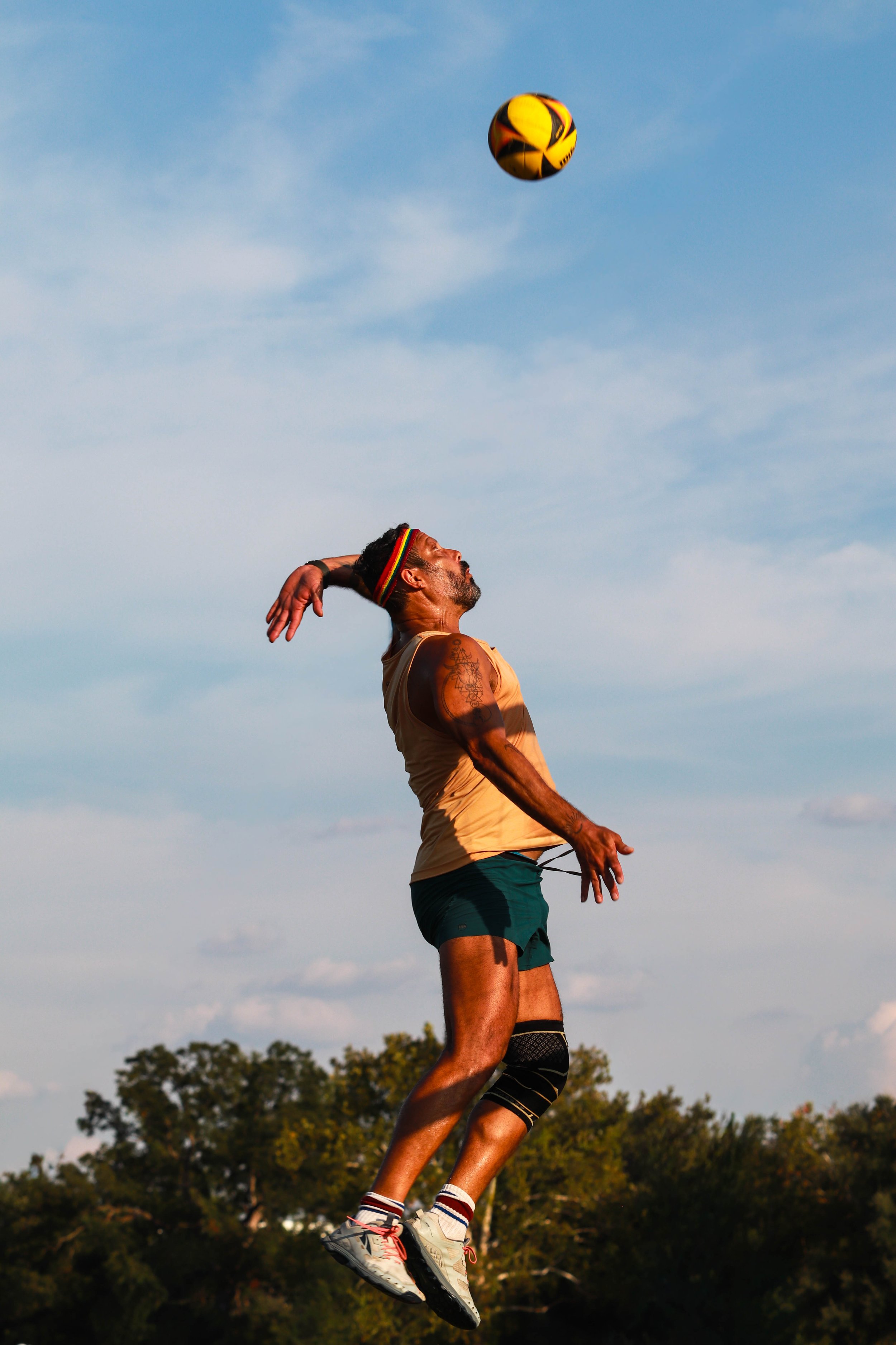 A man with tattoos, wearing a headband, tank top, shorts, and sneakers, is jumping outdoors with a colorful ball in sky, trees in background, under a clear blue sky.
