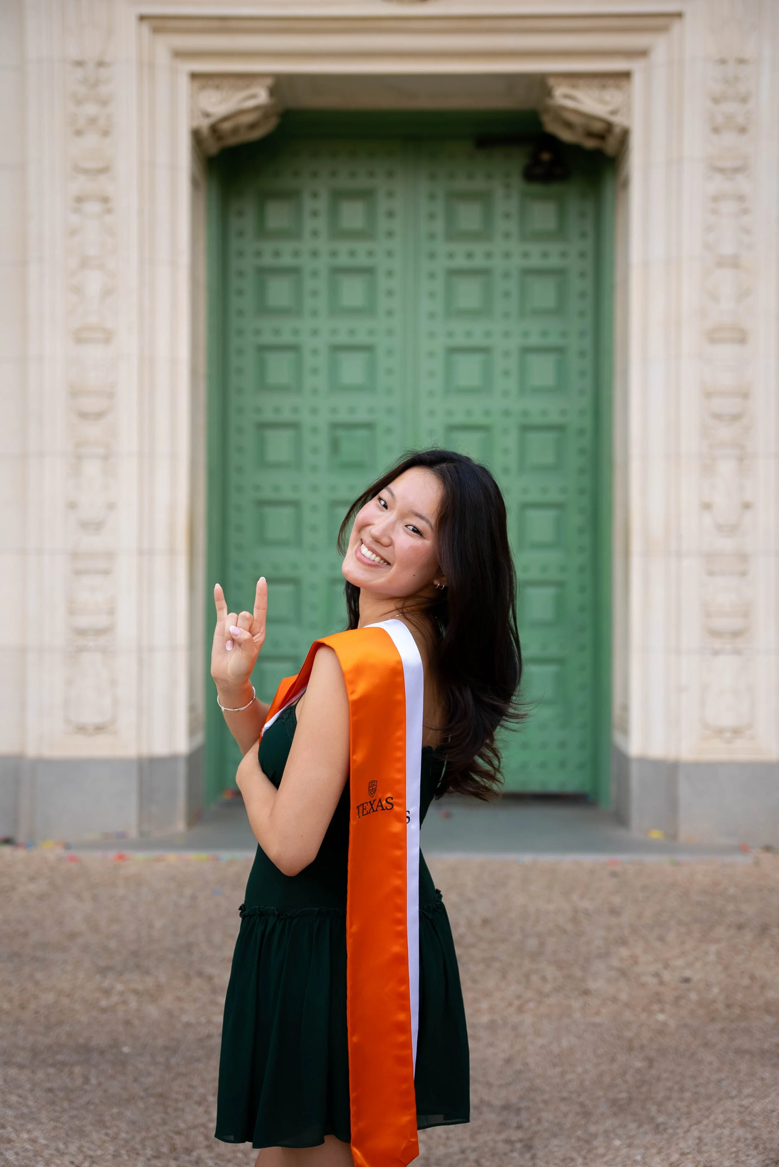 A young woman in graduation attire standing in front of a large green door with light-colored stone framing. She is smiling and making a peace sign with her right hand. She is wearing a dark dress and an orange-and-white sash that says 'TEXAS' on it.