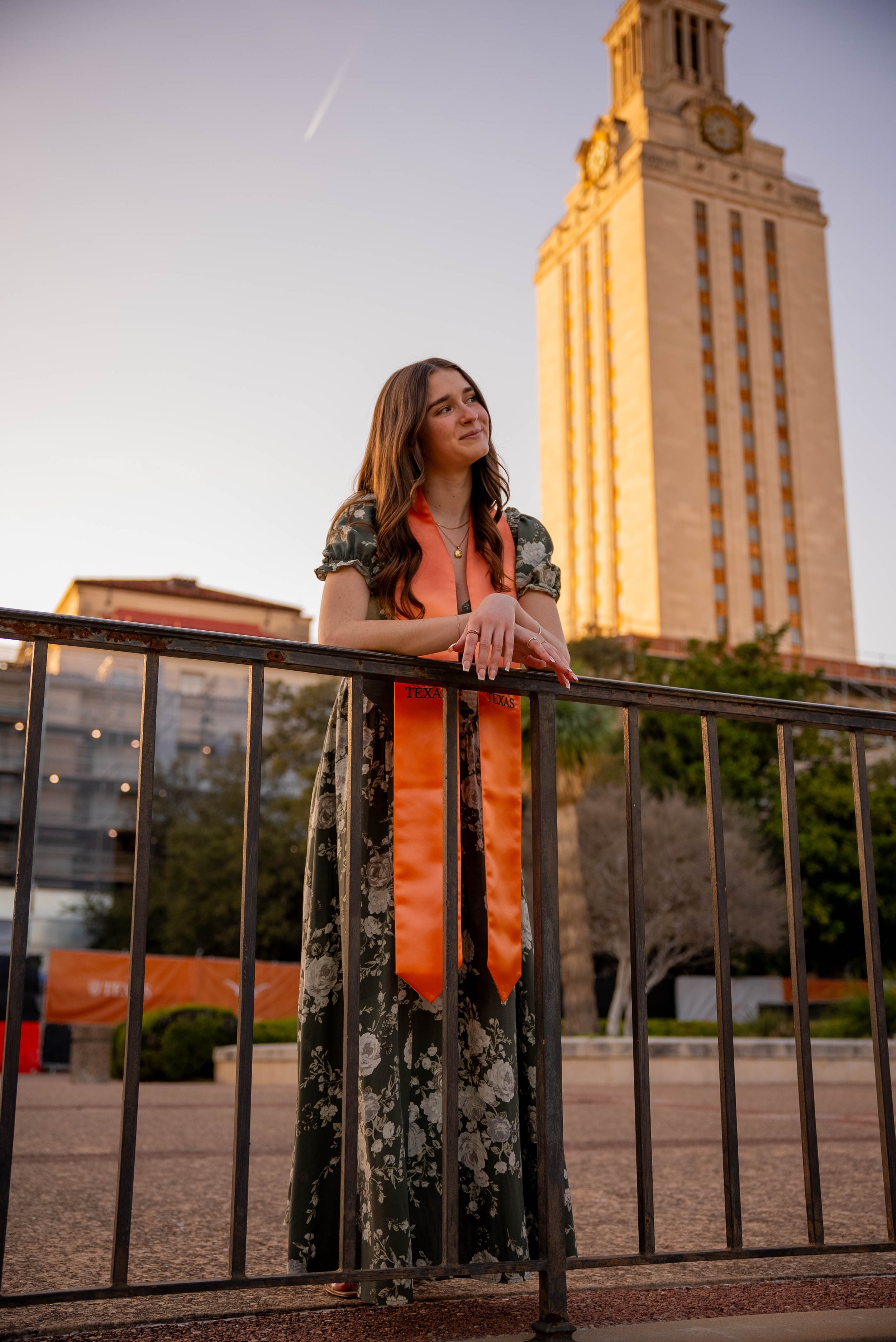 Young woman in floral dress and orange graduation stole leaning on a metal railing in front of a tall building with clocks at sunset.