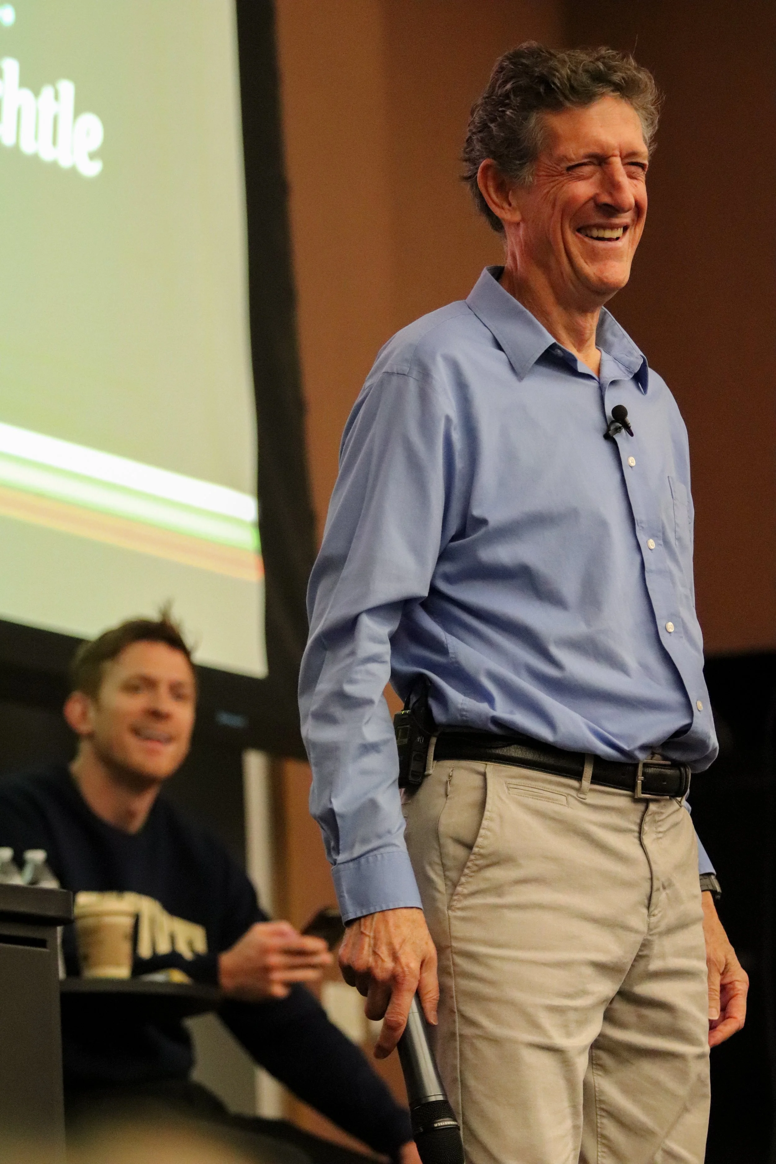 A man with gray hair in a blue shirt is smiling and standing in front of a brown background. Another man with glasses is sitting at a table and looking in the direction of the standing man.